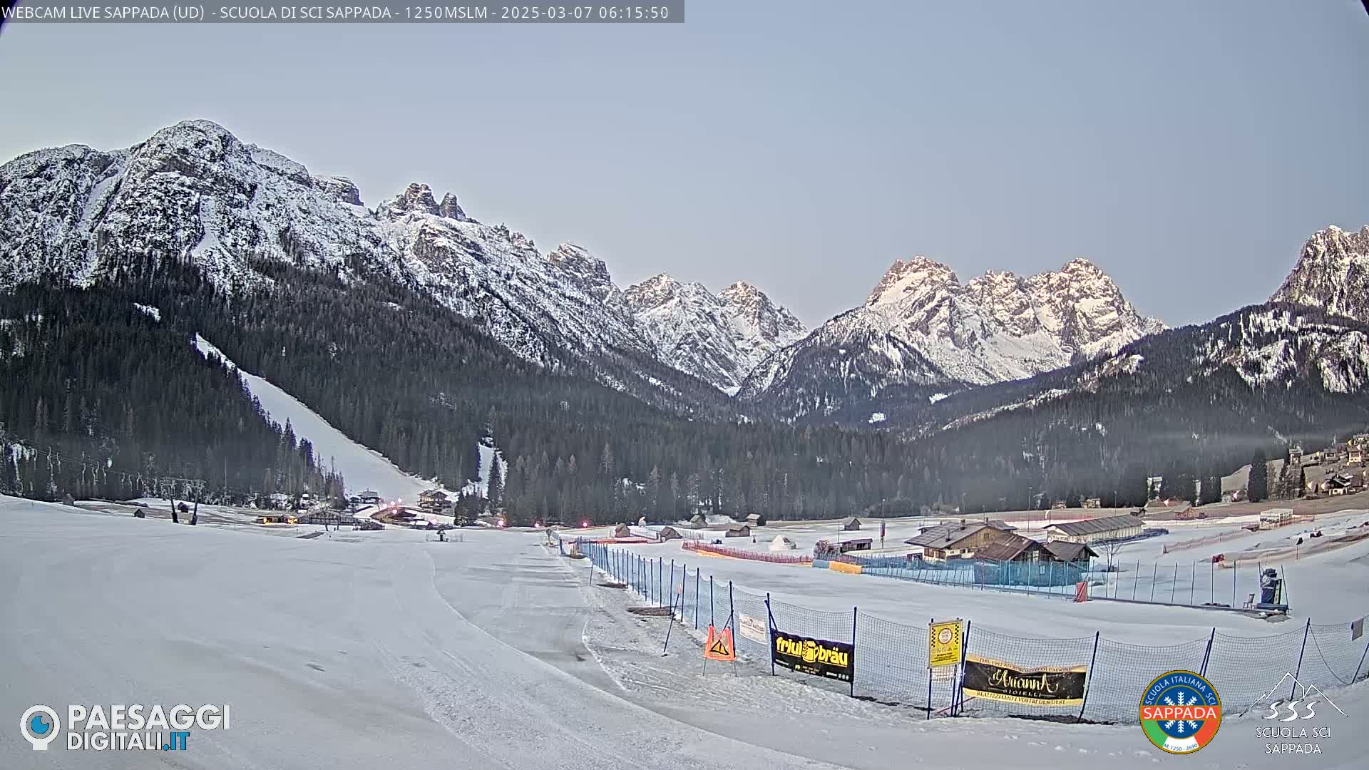 A snow-covered ski slope and surrounding valley are nestled beneath a range of snow-capped mountains under a clear, twilight sky.