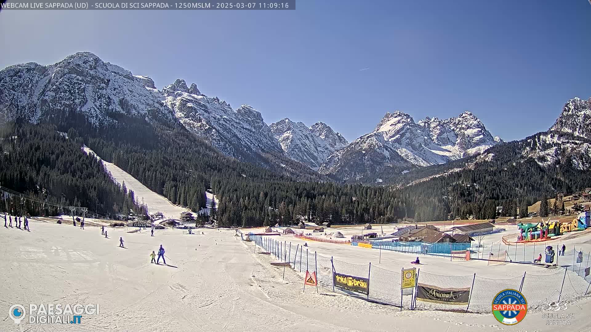 A sunny day on a snow-covered ski slope with a few skiers and a backdrop of snow-capped mountains.