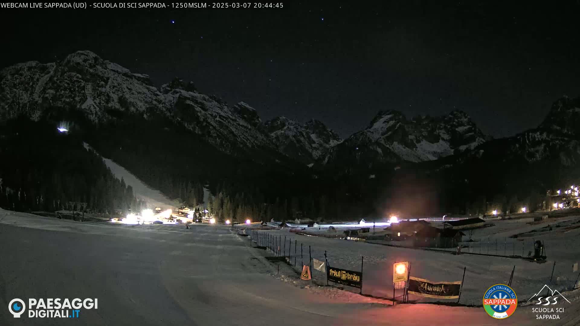 A nighttime view of a snow-covered ski slope at the base of dark mountains under a clear, starry sky.