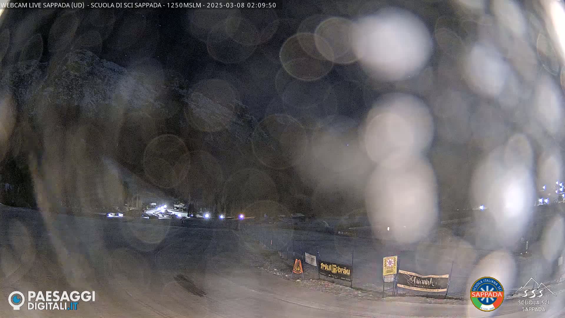 A nighttime view of a snow-covered area with some lights visible in the distance, under a dark, possibly hazy sky.