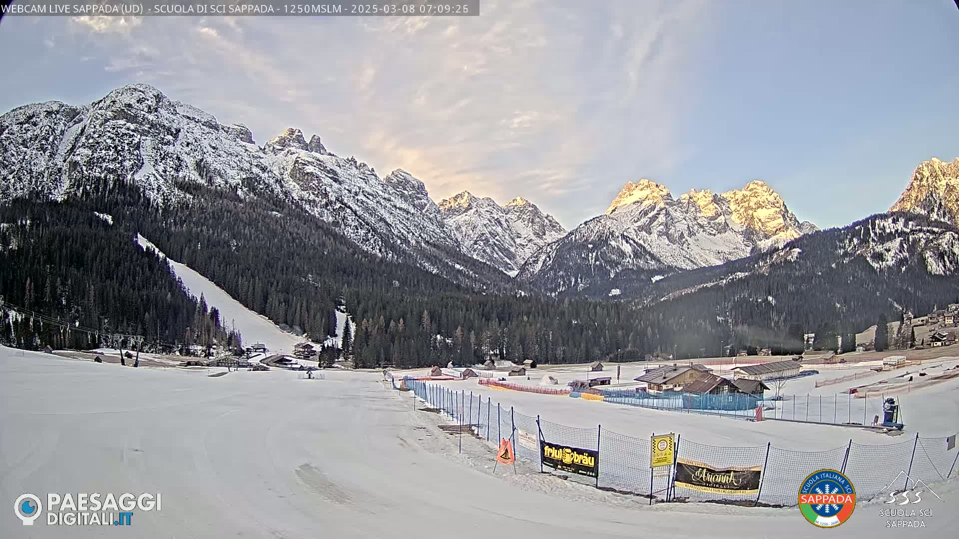 A snow-covered ski slope and surrounding snow-capped mountains are visible under a partly cloudy sky at sunrise.