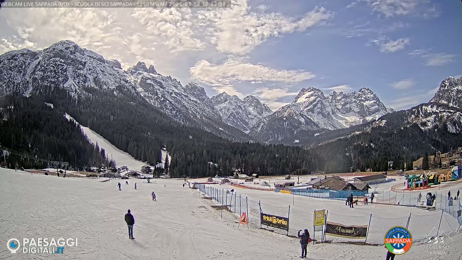 A snow-covered ski slope with several people skiing and snowboarding, situated in a valley surrounded by snow-capped mountains under a partly cloudy sky.