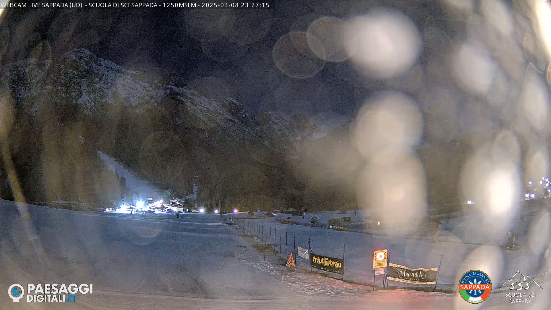 A nighttime view of a snow-covered ski slope at the base of a snow-dusted mountain range, under a hazy, dark sky.