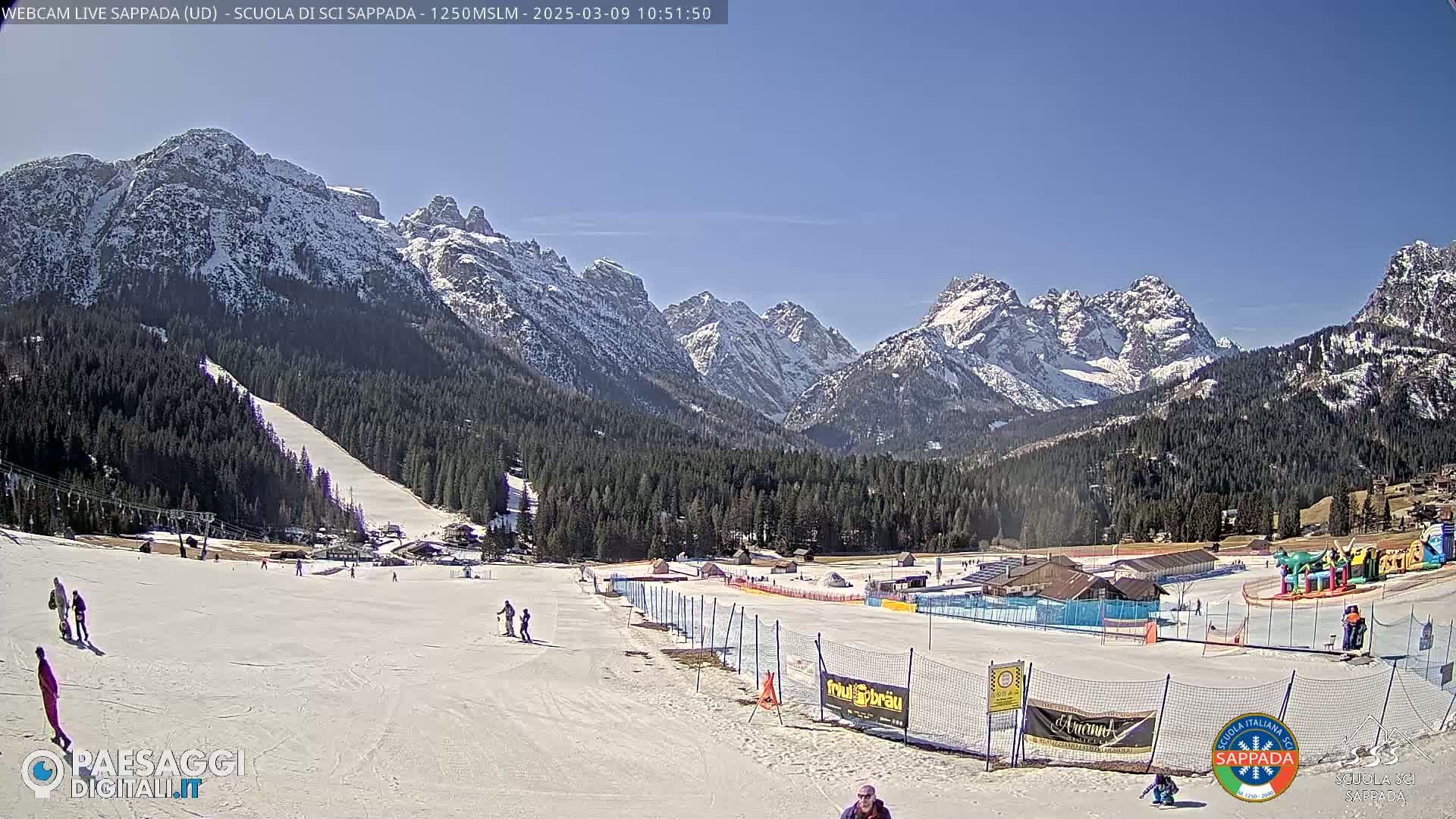 A sunny day at a snow-covered ski resort in the mountains, showing several people skiing and snowboarding on a gently sloping piste.