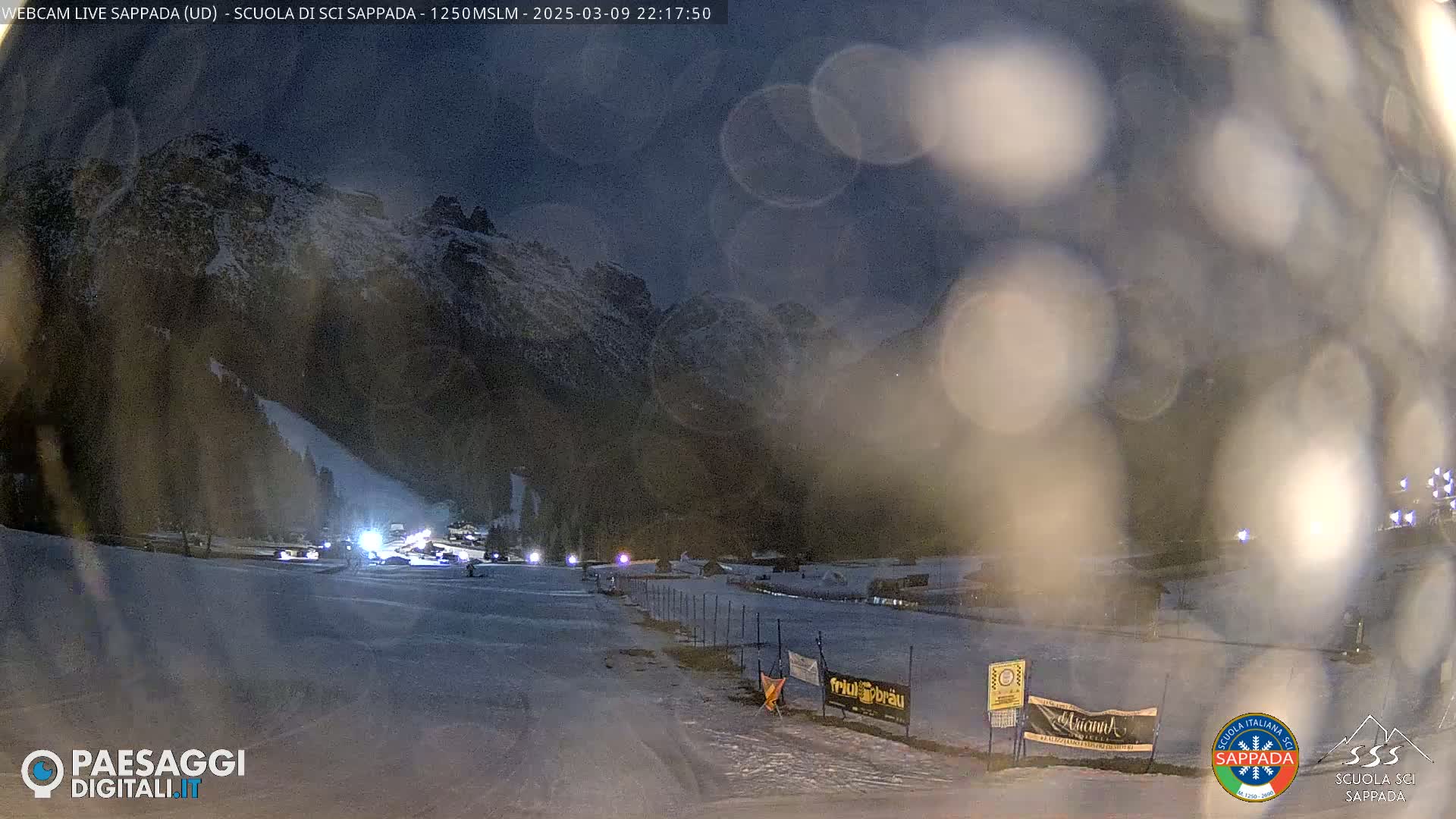A nighttime, snowy scene shows a ski slope with a few vehicles and lights at the base, under a dark sky with some snow falling and a mountain in the background.