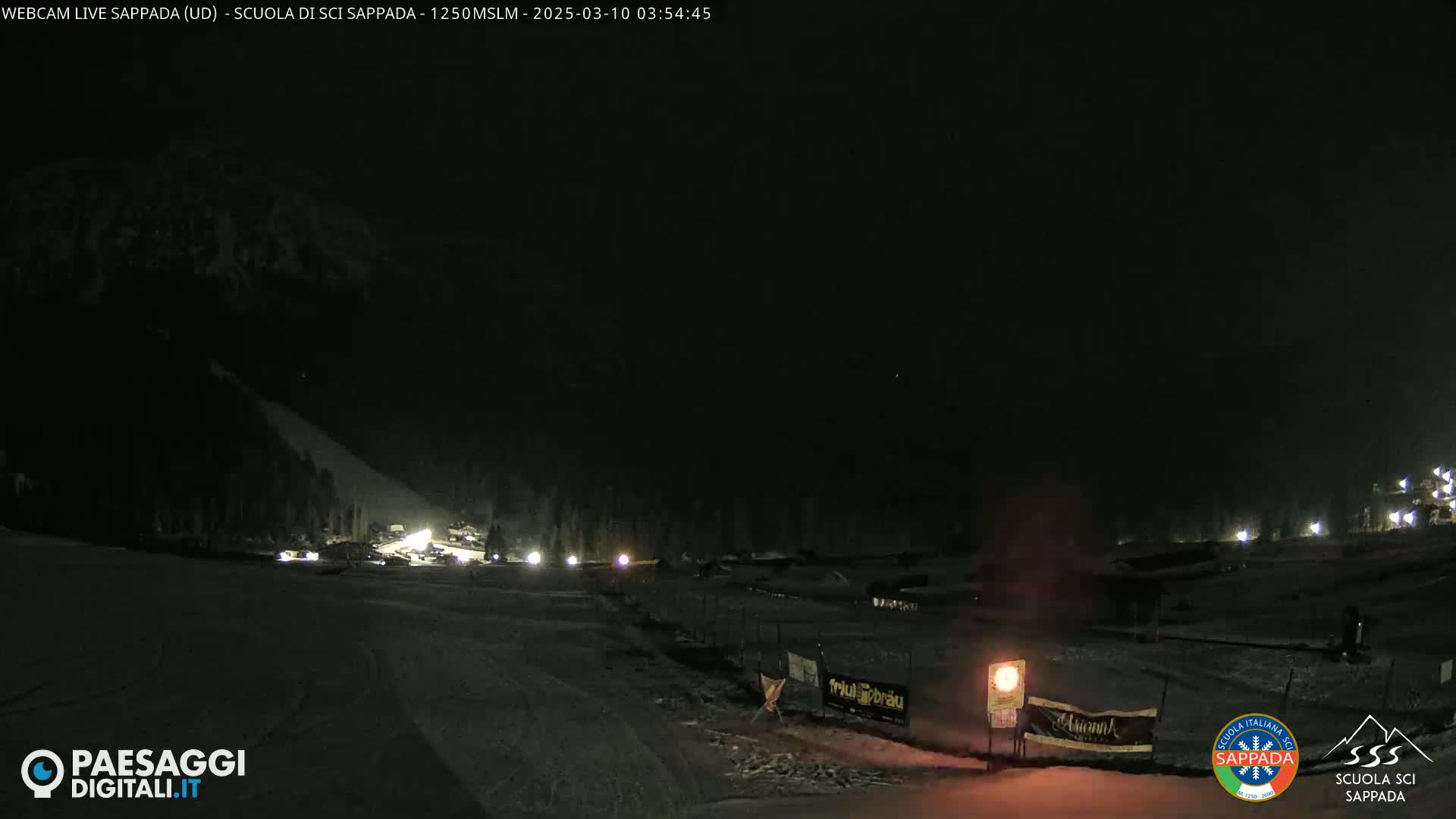 A nighttime view of a snow-covered ski slope with dimly lit buildings and sparsely visible vehicles.