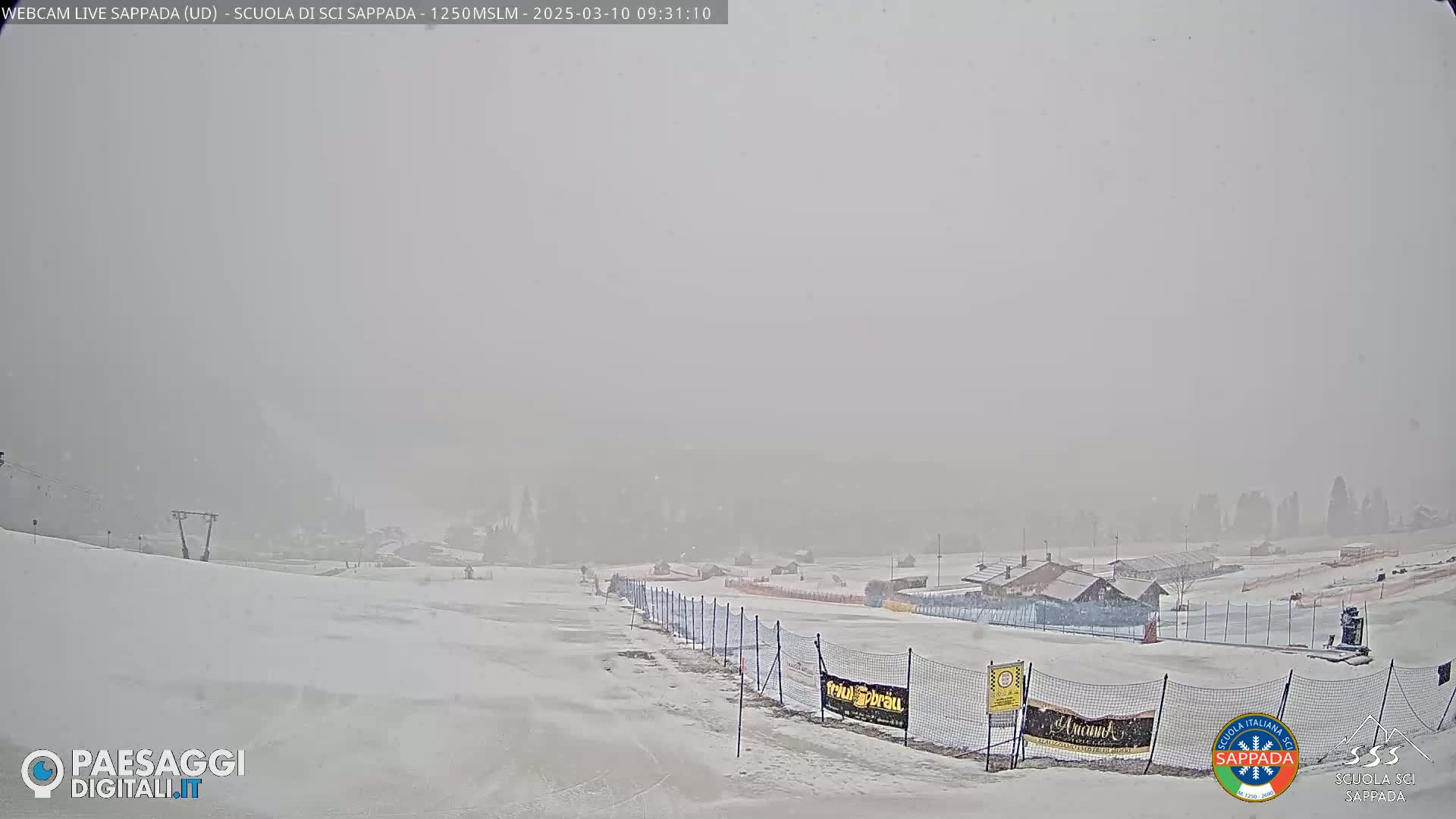 A snow-covered ski slope is visible through a heavy snowstorm, with buildings and ski lifts barely discernible in the distance.