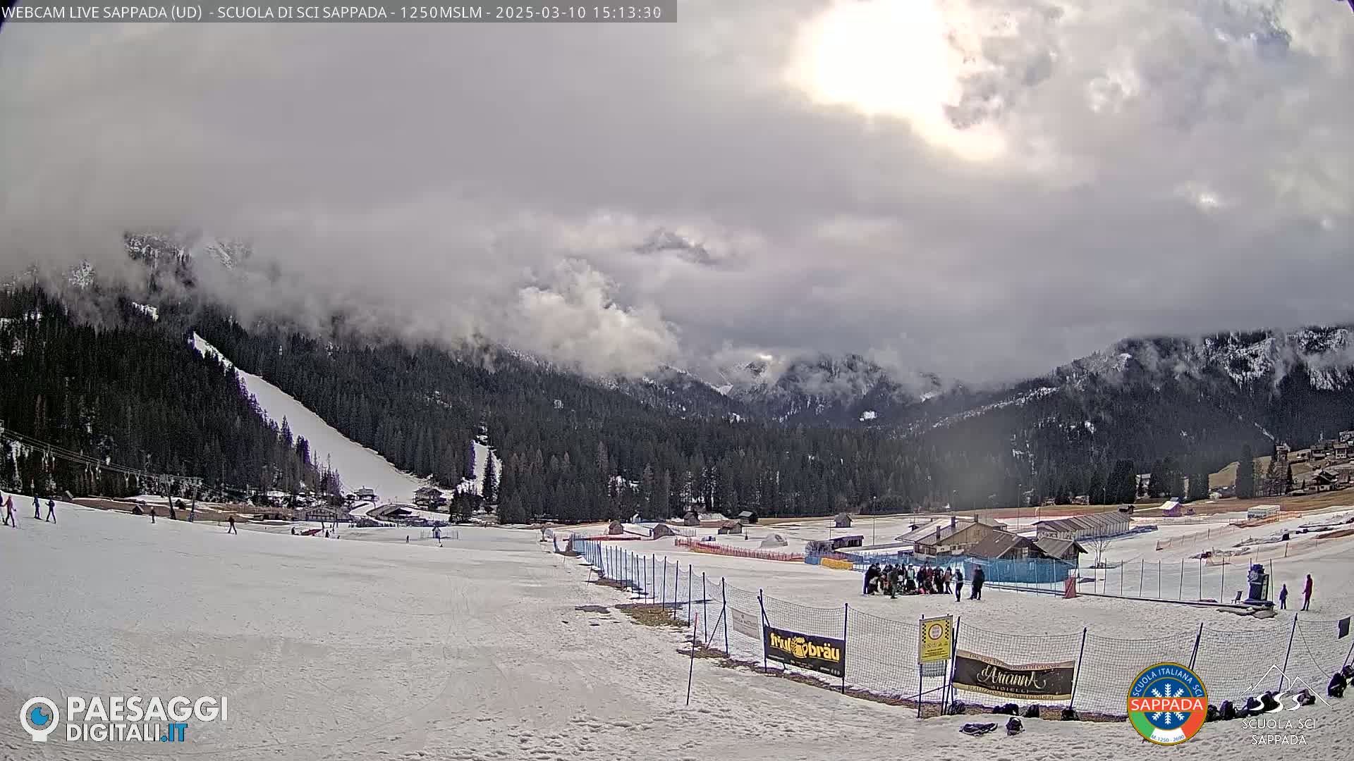 A snowy ski slope with a few skiers and a mountain range in the background under a mostly cloudy sky.