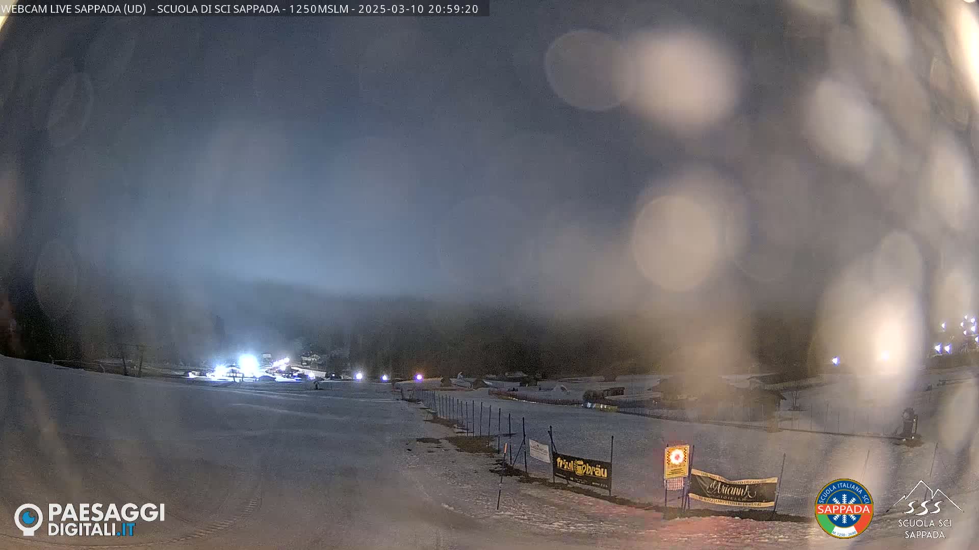 A nighttime view of a snow-covered ski slope with some lights visible in the distance, under a hazy, dark sky.