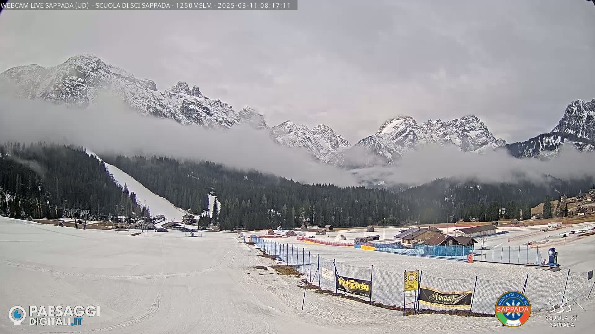 A snowy ski slope at the base of fog-shrouded, snow-capped mountains under an overcast sky.