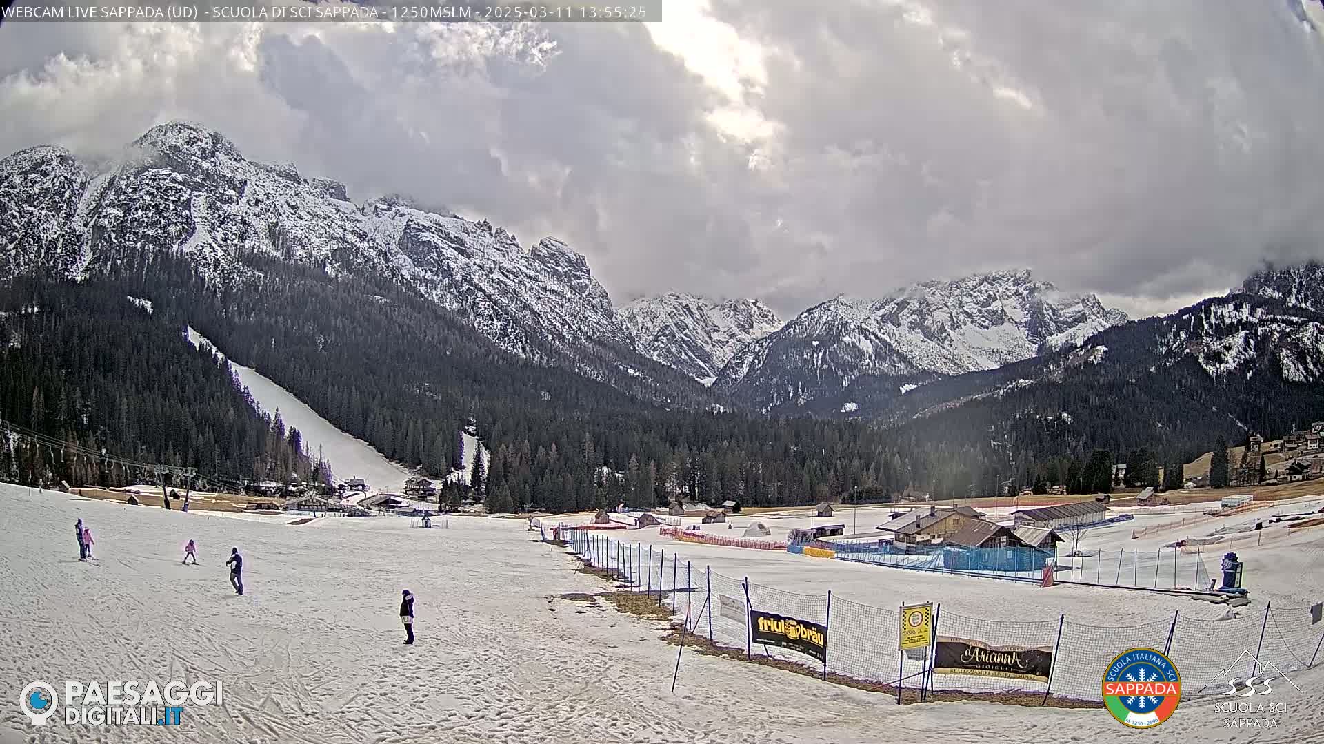 A mostly cloudy day reveals a snow-covered ski slope at the foot of snow-capped mountains, with a few skiers visible.