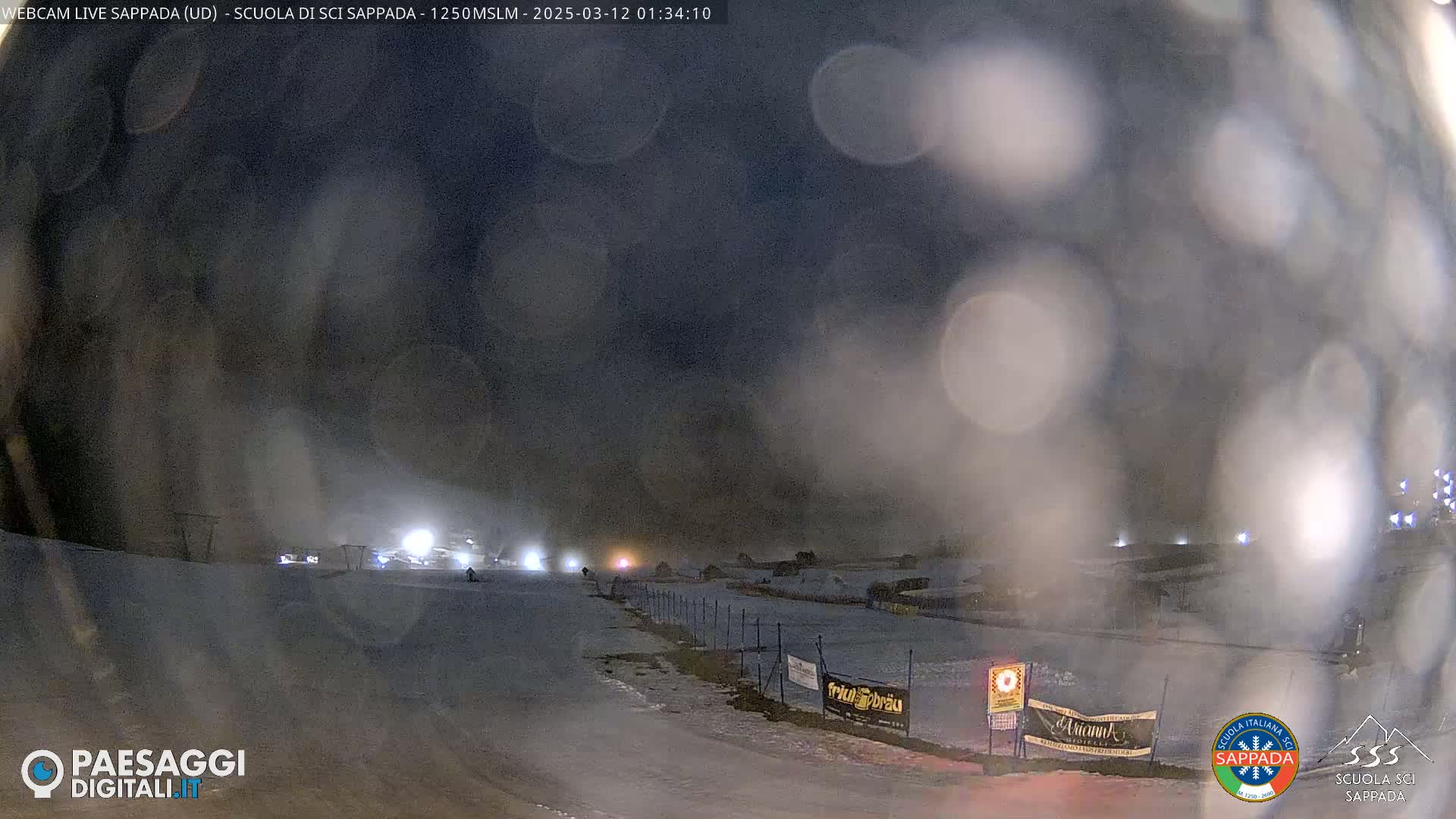 A snowy nighttime scene at a ski resort, with lights illuminating a slope and a fence in the foreground, under hazy conditions.