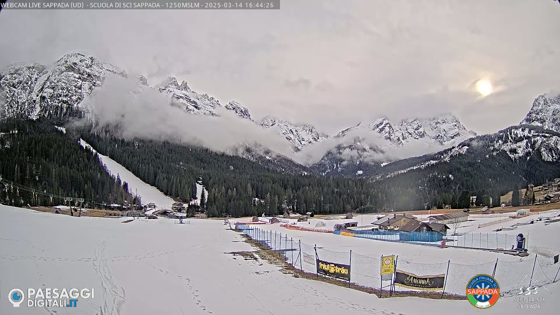 A snow-covered ski slope and valley are nestled beneath partially cloudy skies and snow-capped mountains, with a partially visible sun.