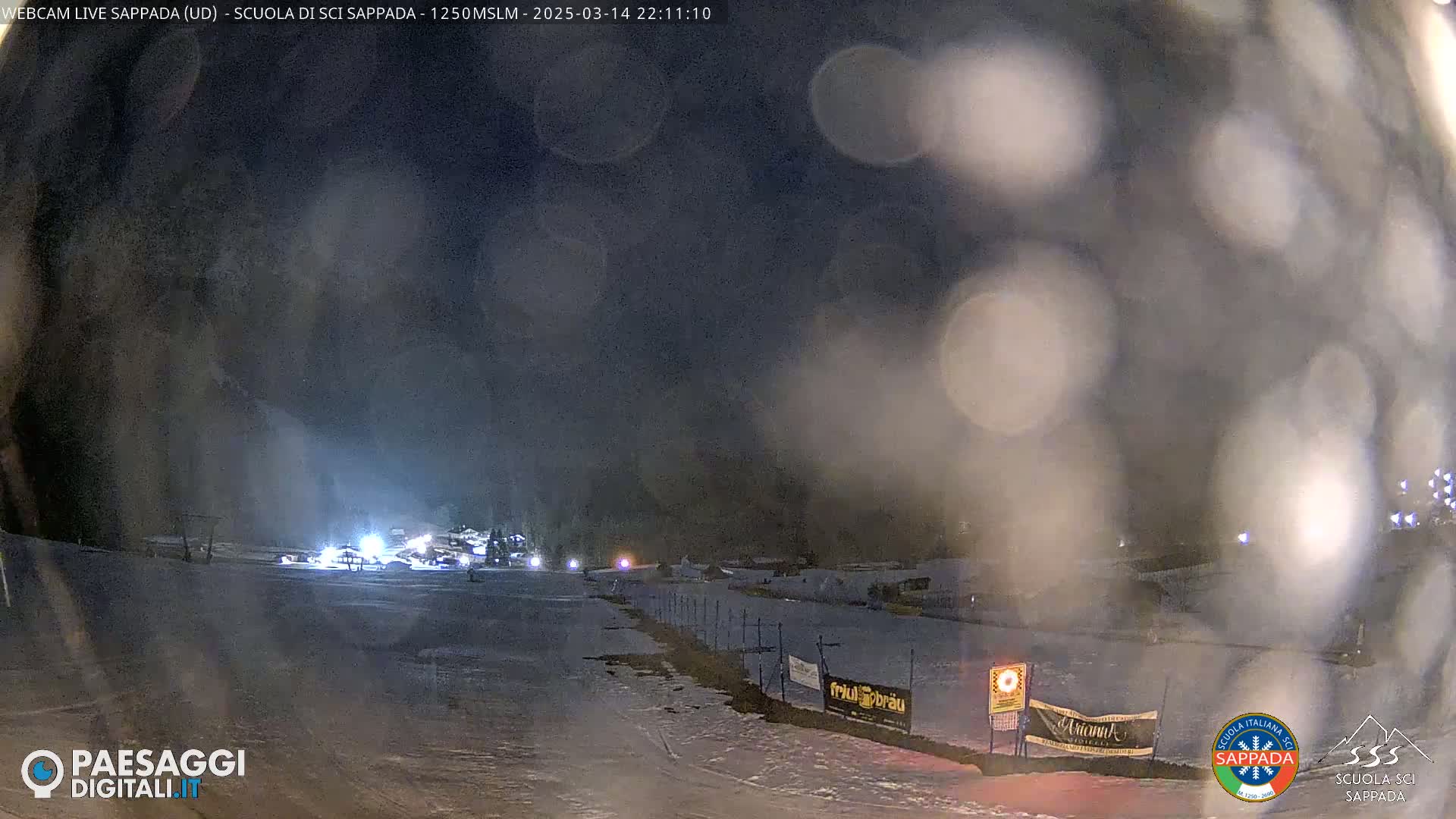A nighttime snow-covered ski slope is dimly lit, with a few lights visible in the distance, under a hazy, dark sky.