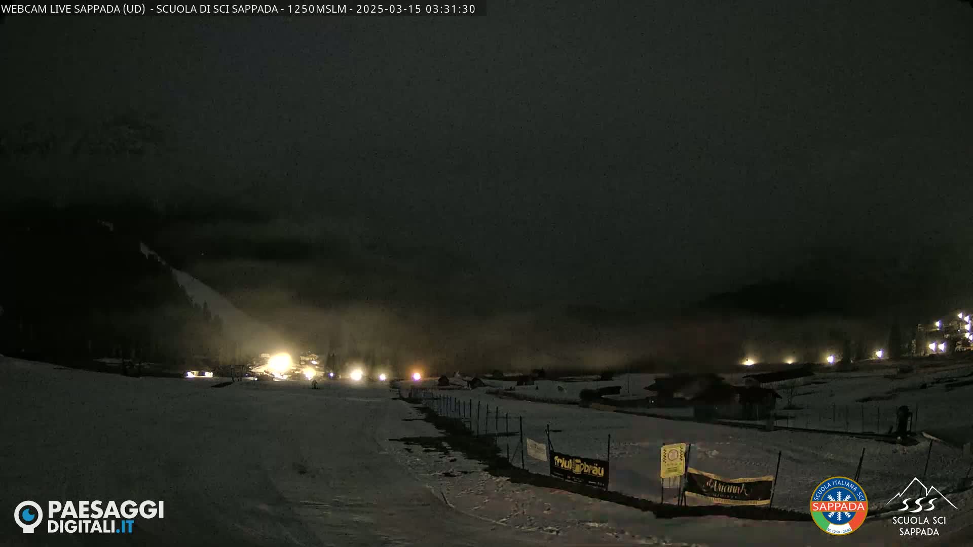 A nighttime view of a snow-covered ski slope with low fog or clouds, illuminated by scattered lights.