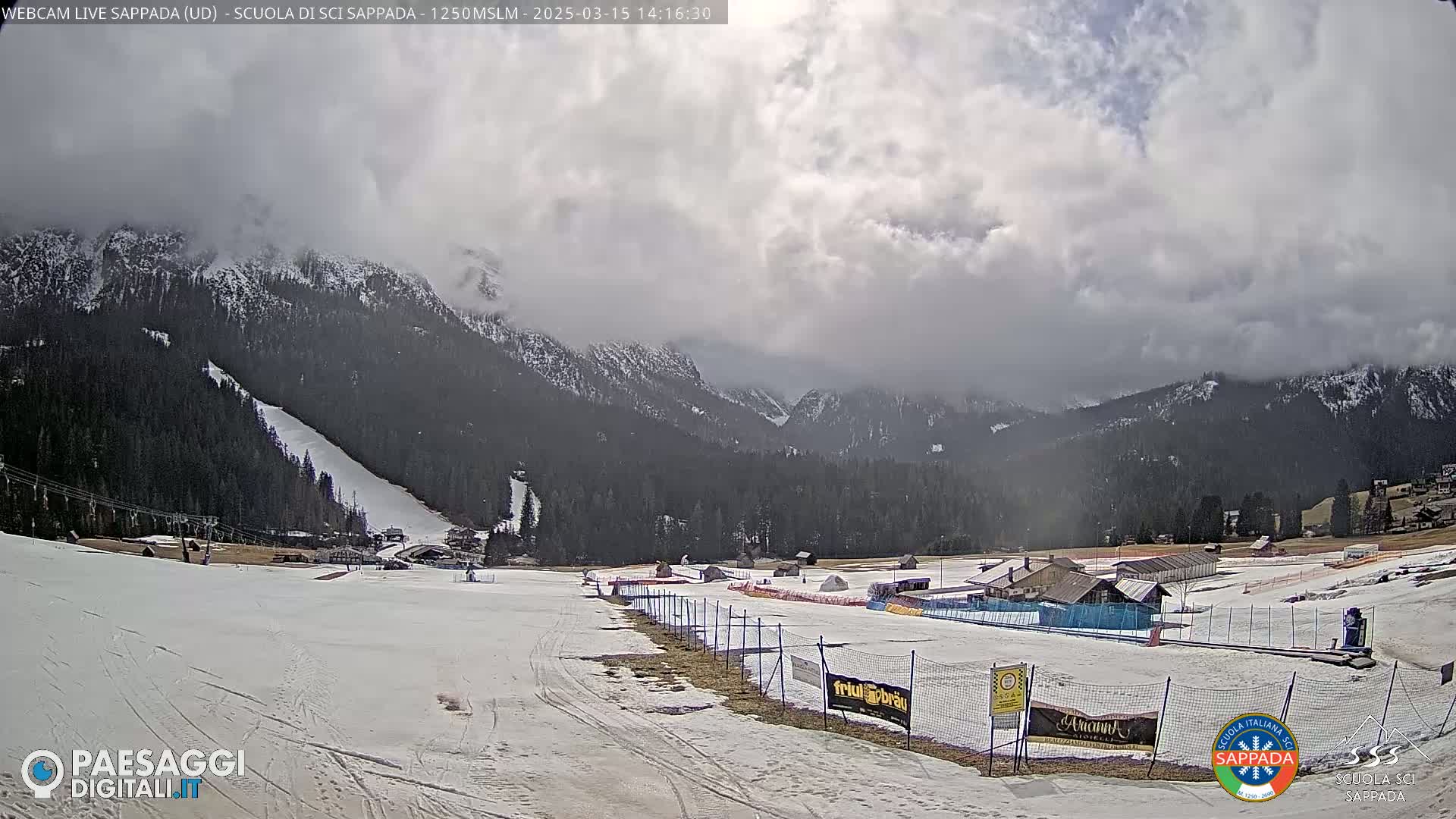 A partly cloudy day reveals a snow-covered ski slope nestled in a valley between snow-capped mountains, with several buildings and fences visible in the foreground.