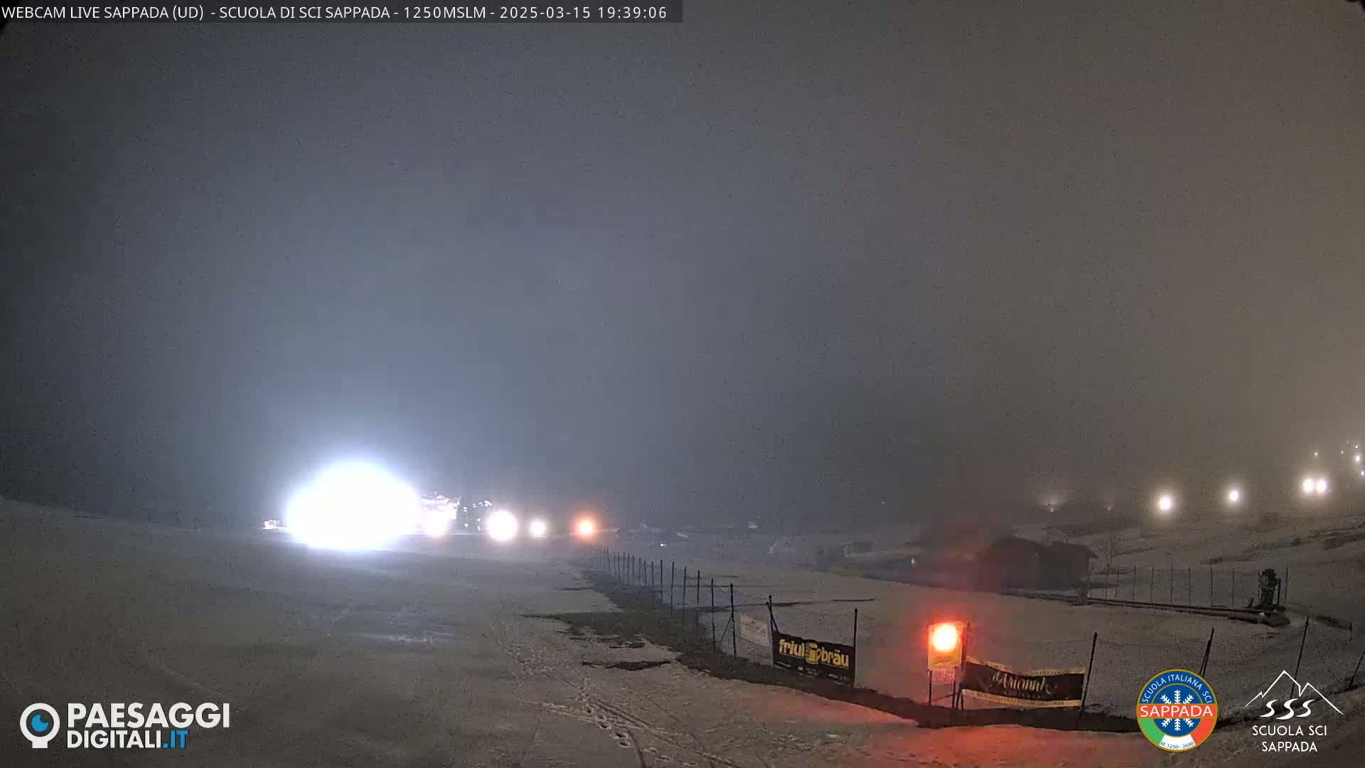 A snowy, foggy nighttime scene shows a dimly lit area with a fence, some buildings, and bright lights in the distance.