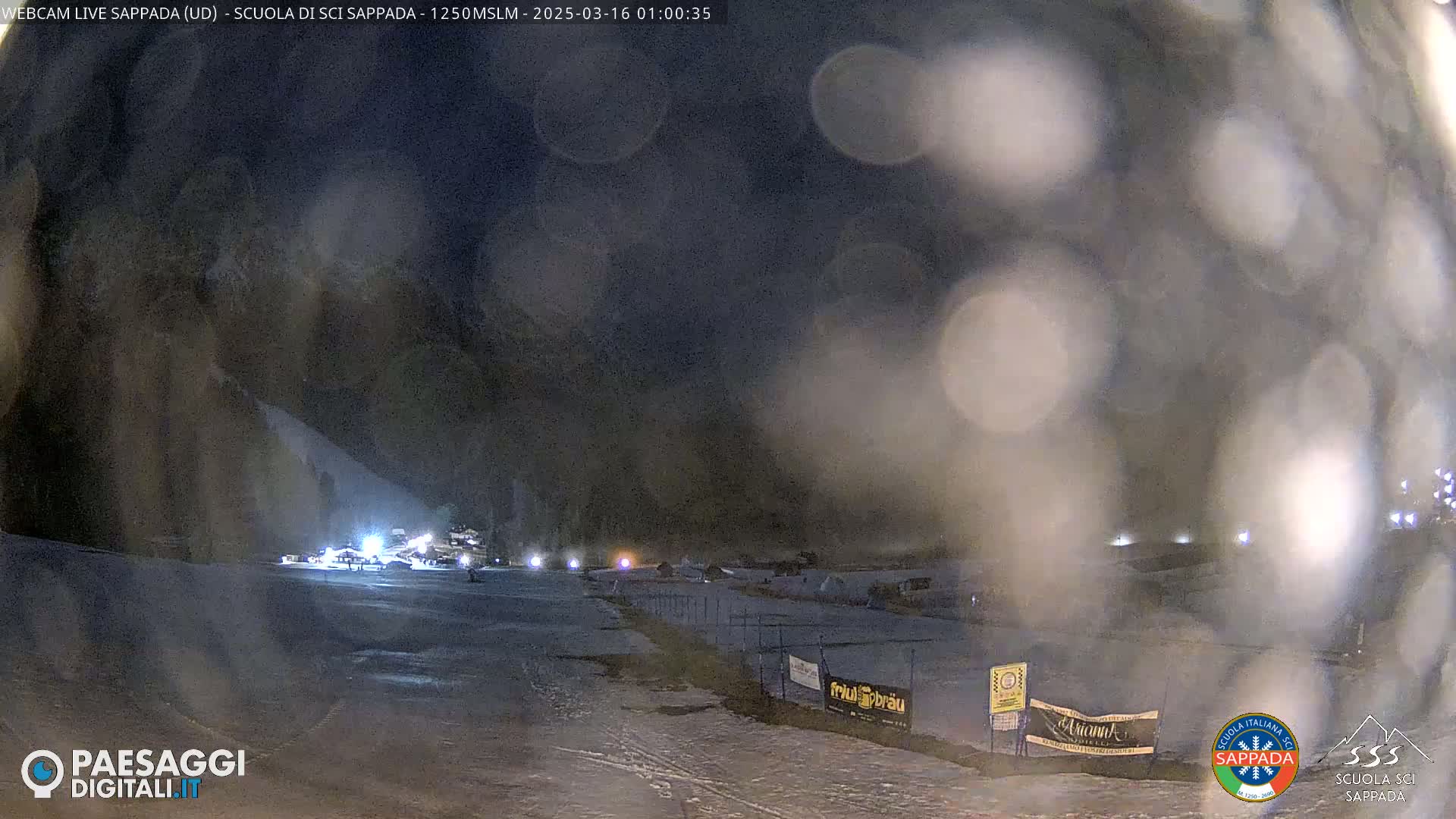 A nighttime view of a snow-covered ski slope with some lights and figures visible in the distance, obscured by a layer of what appears to be rain or snow on the camera lens.