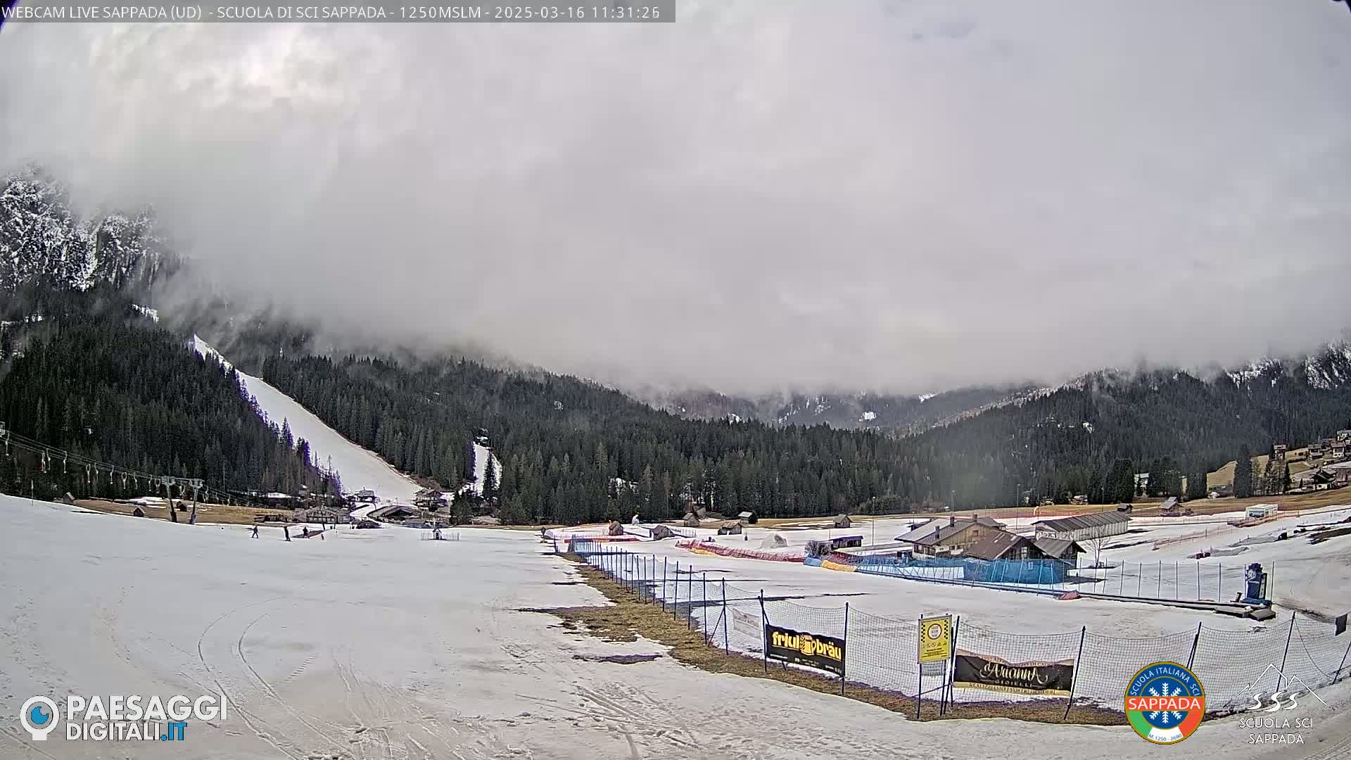 A snowy ski slope with a few skiers is visible beneath a cloudy sky, overlooking a mountain range partially obscured by fog.