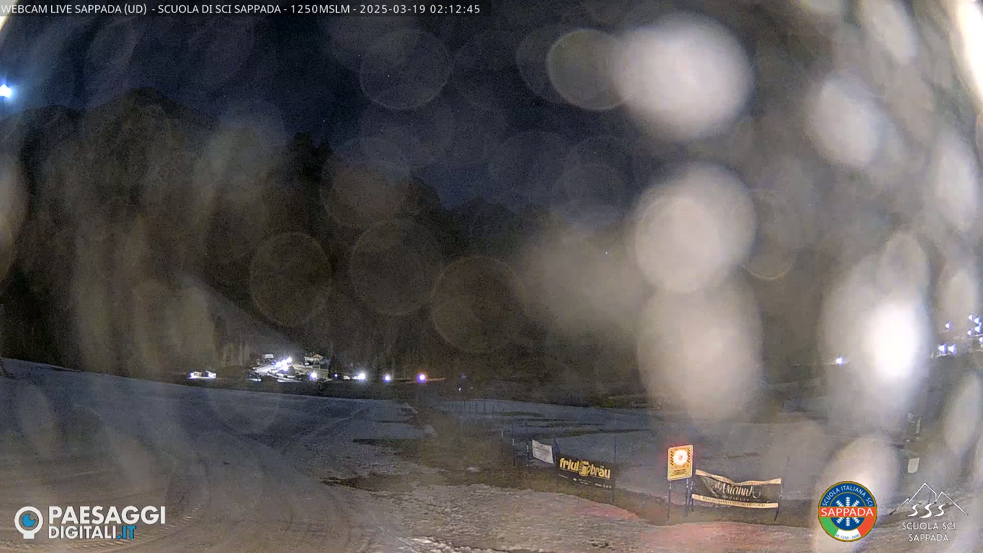 A nighttime, snowy scene is viewed through a blurry lens, showing a dimly lit area with some vehicles and fencing.