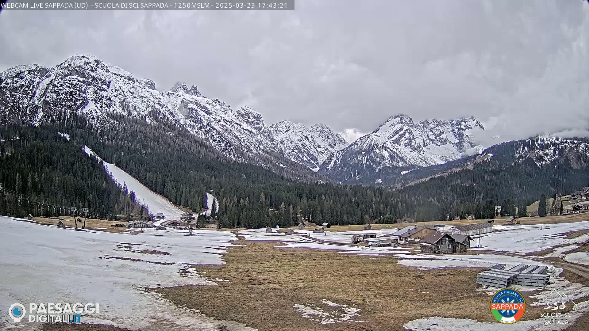 A partially cloudy day shows a snow-dusted mountain range overlooking a valley with patchy snow cover, a ski slope, and several buildings.