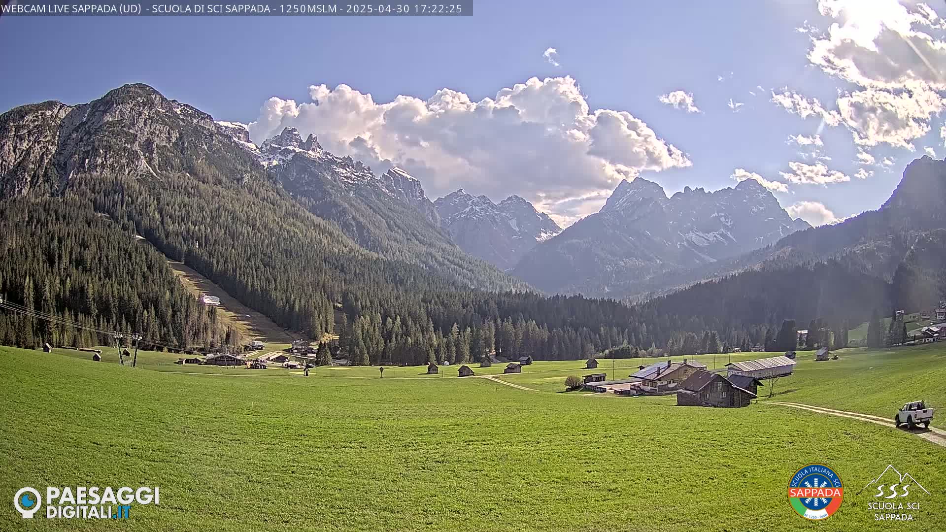 A partly cloudy day reveals a valley of green fields and small buildings nestled amongst a dark forest, with snow-capped mountains rising in the background.