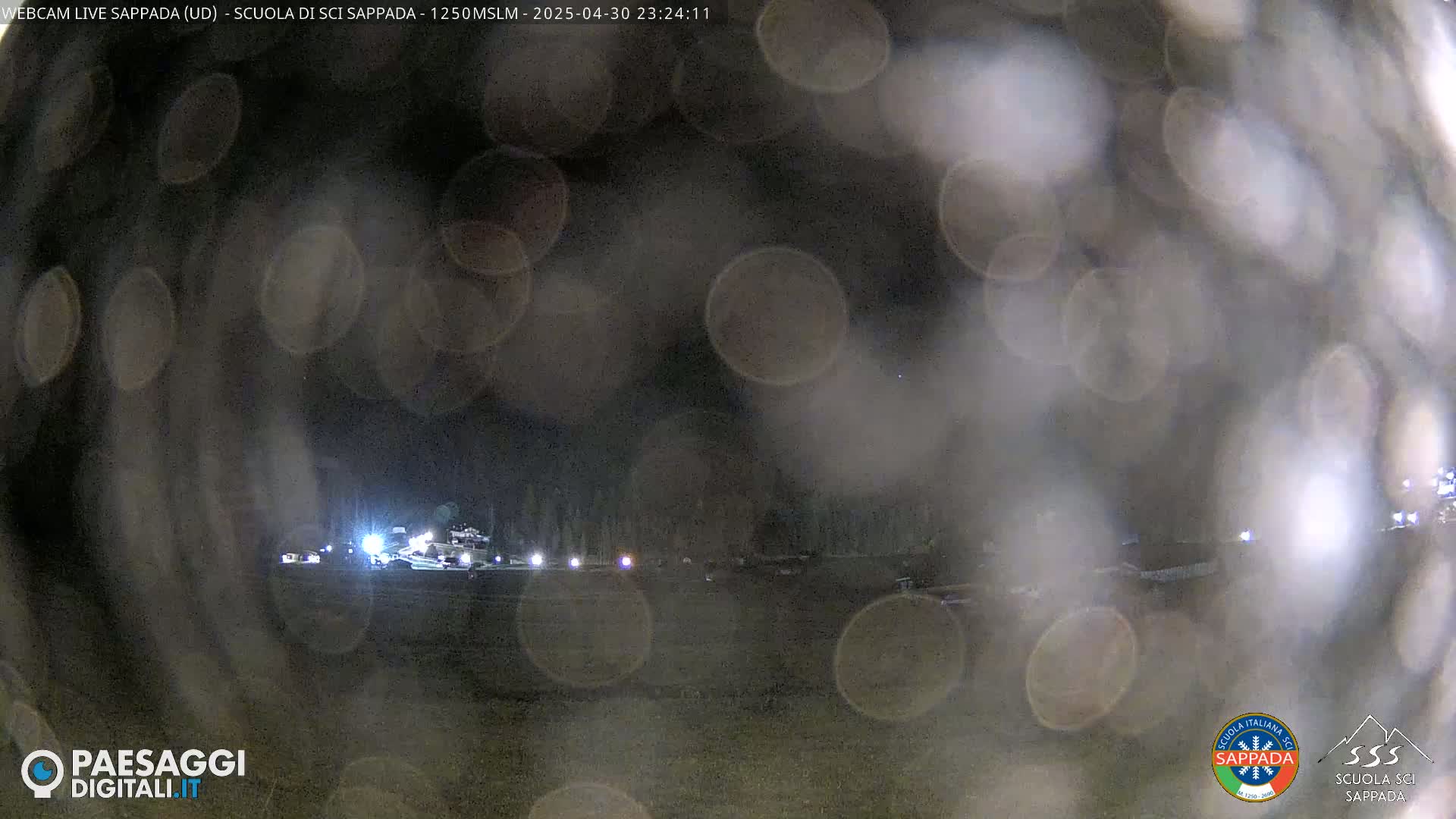 A nighttime view of a snow-covered area with some lights visible in the distance through blurry, rain-covered glass.