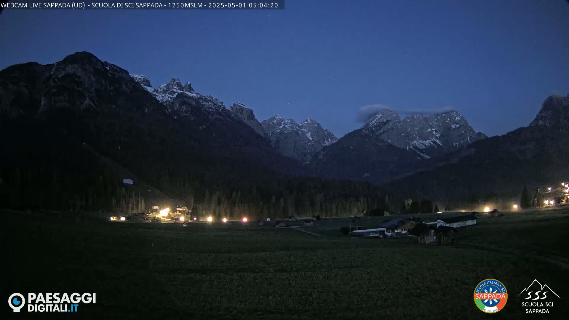 Snow-capped mountains are silhouetted against a twilight sky over a dark, flat valley with a few scattered lights.