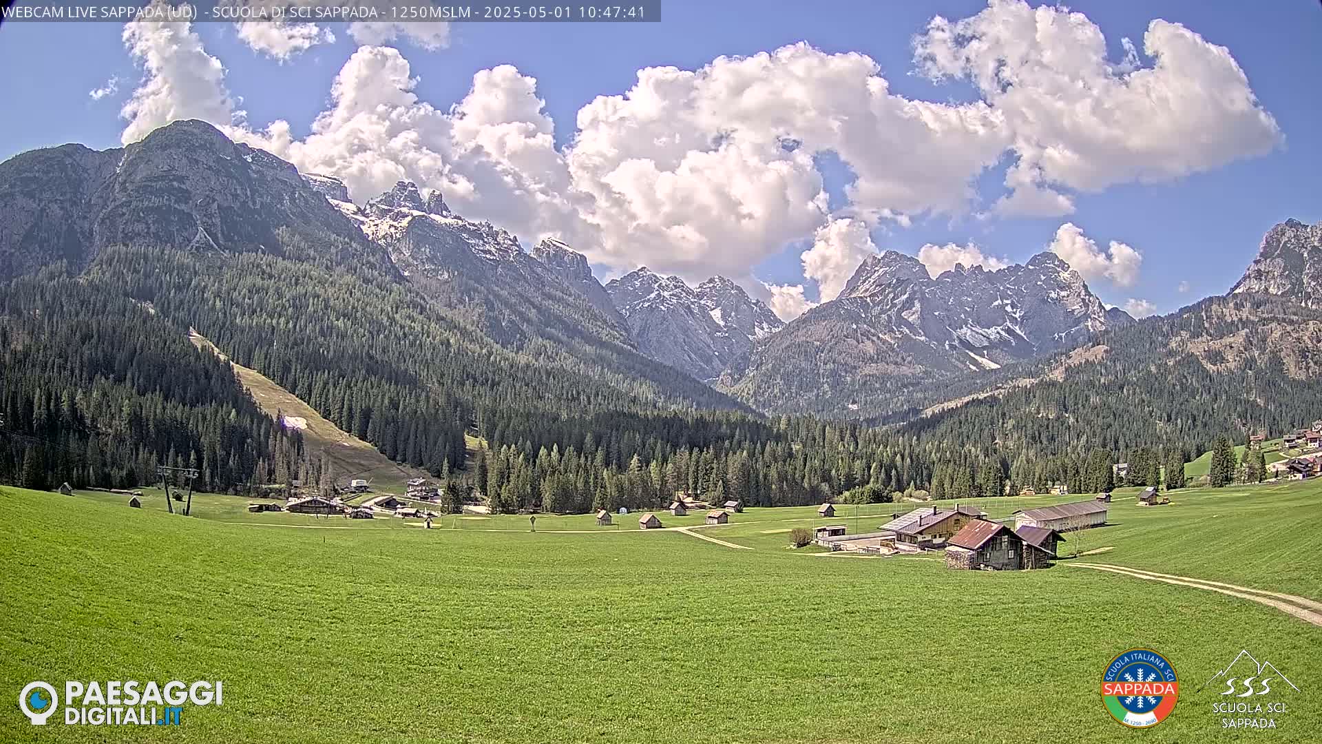 A partly cloudy day reveals a verdant valley with scattered buildings, a ski slope, and a backdrop of snow-capped mountains.