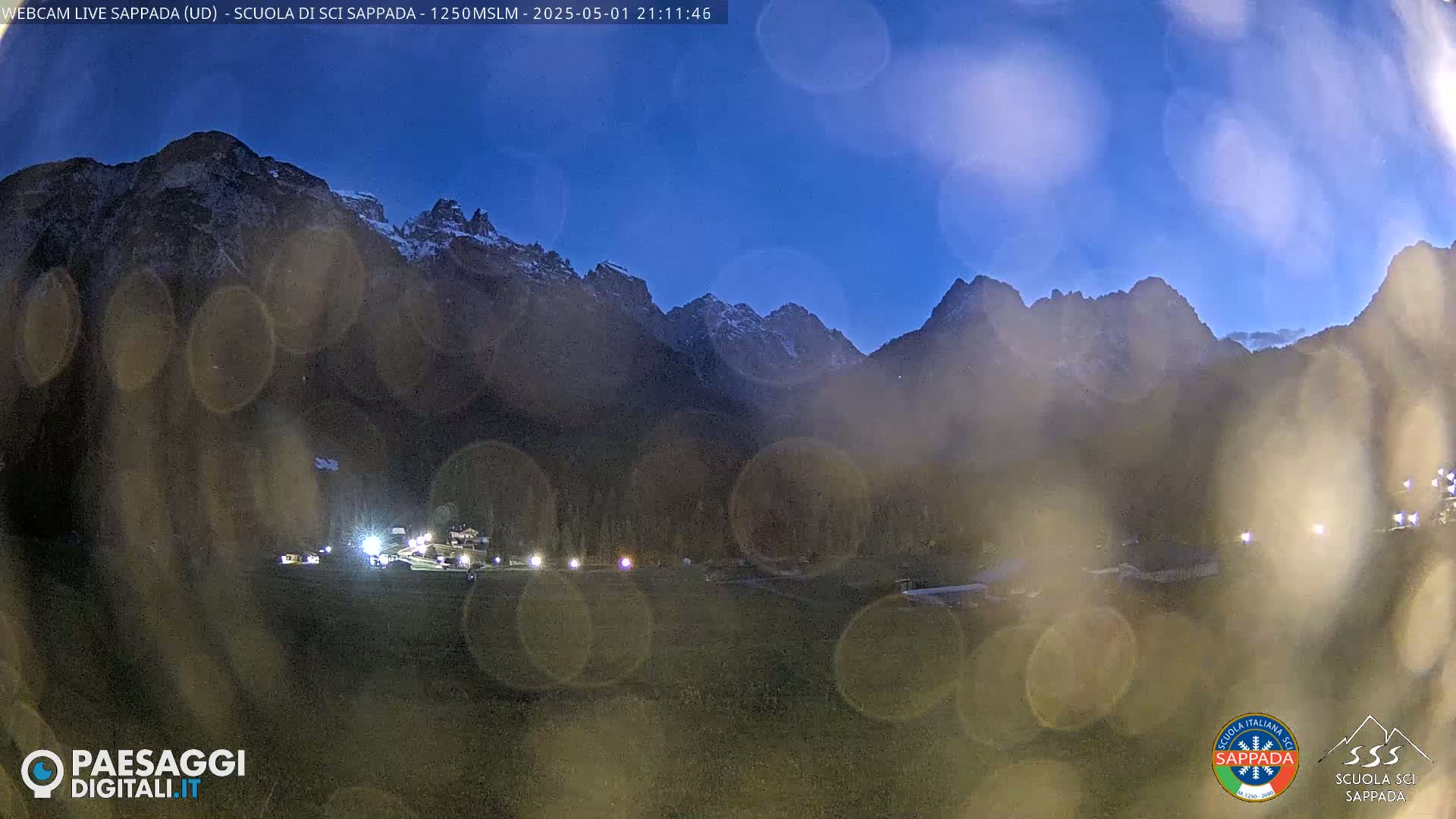 A nighttime view of snow-capped mountains, seen through rain-streaked glass, with a small village illuminated in the foreground under a twilight sky.
