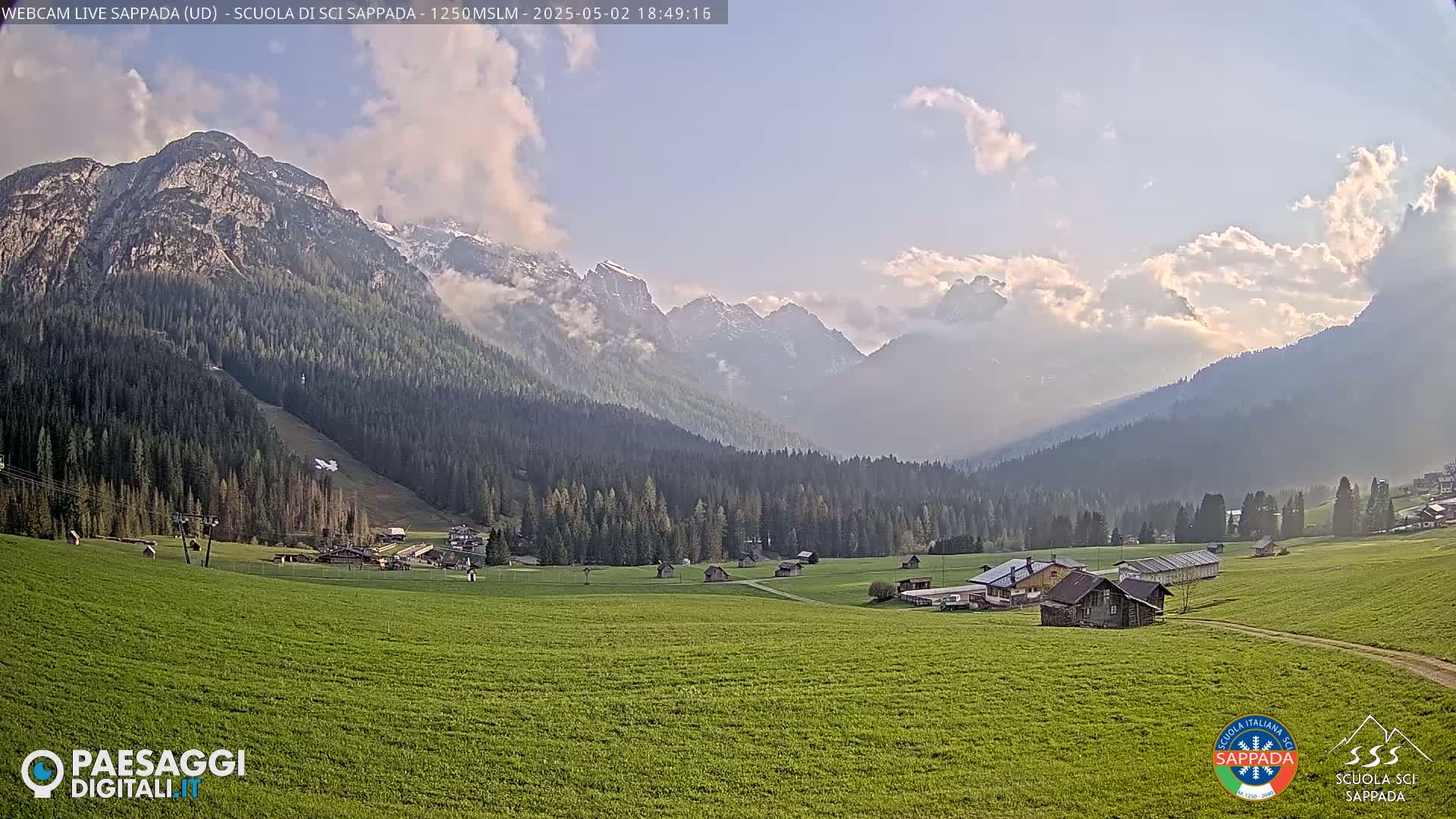 A valley of green fields and a few buildings is nestled beneath a mountain range partially covered in snow and clouds under a partly cloudy sky.