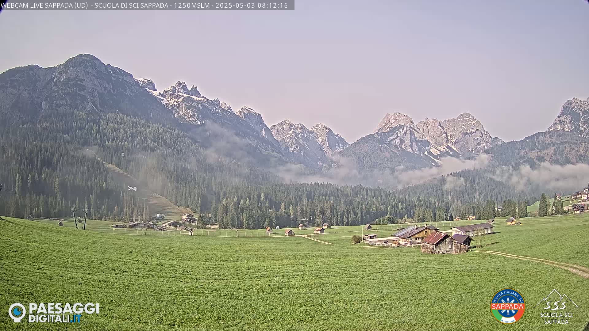 A green valley dotted with small buildings is nestled beneath a mountain range partially obscured by fog, with patches of snow visible on the higher peaks.