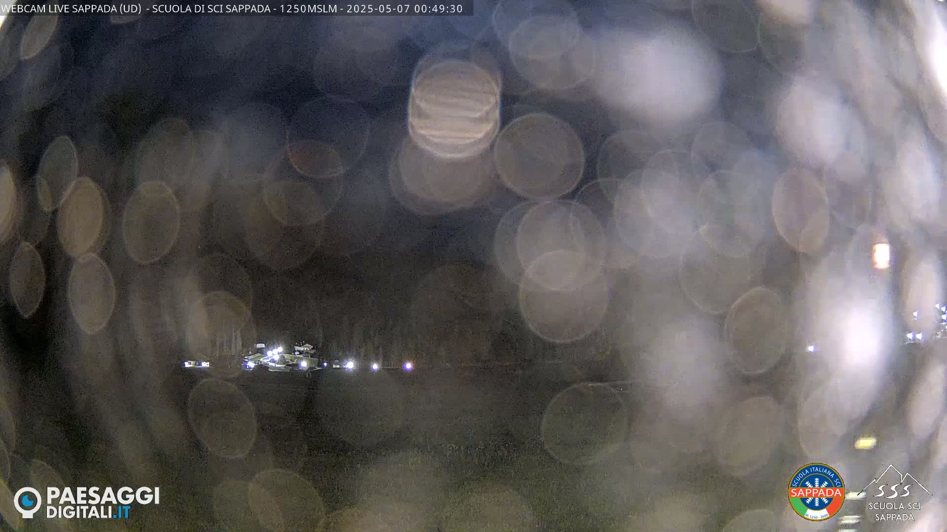 A nighttime, blurry view of a snow-covered landscape with lights in the distance, obscured by rain or snow on the camera lens.