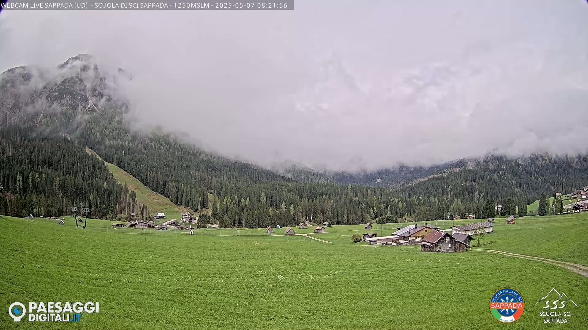 A misty mountain valley with a green field, several small buildings, and a forest in the background.