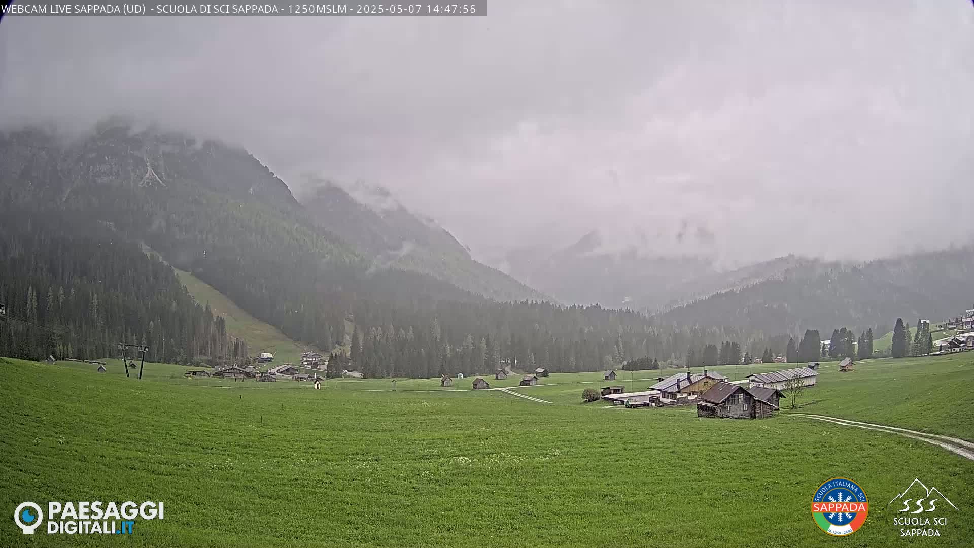 A misty, overcast view shows a valley of green fields and small buildings nestled among dark green forests and imposing mountains.