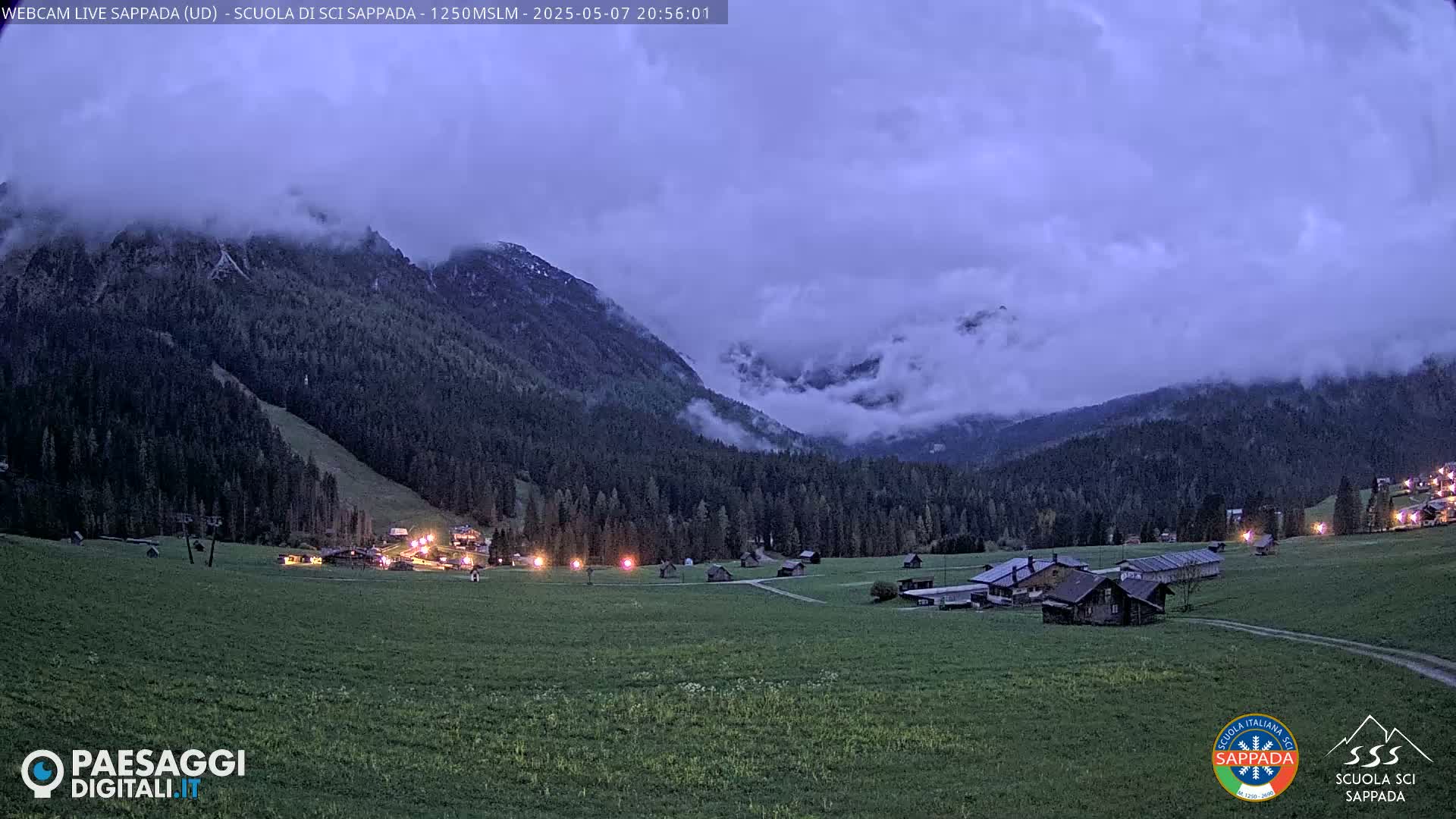 A valley with a few small buildings and a grassy field is nestled beneath mountains shrouded in low-hanging clouds under a twilight sky.