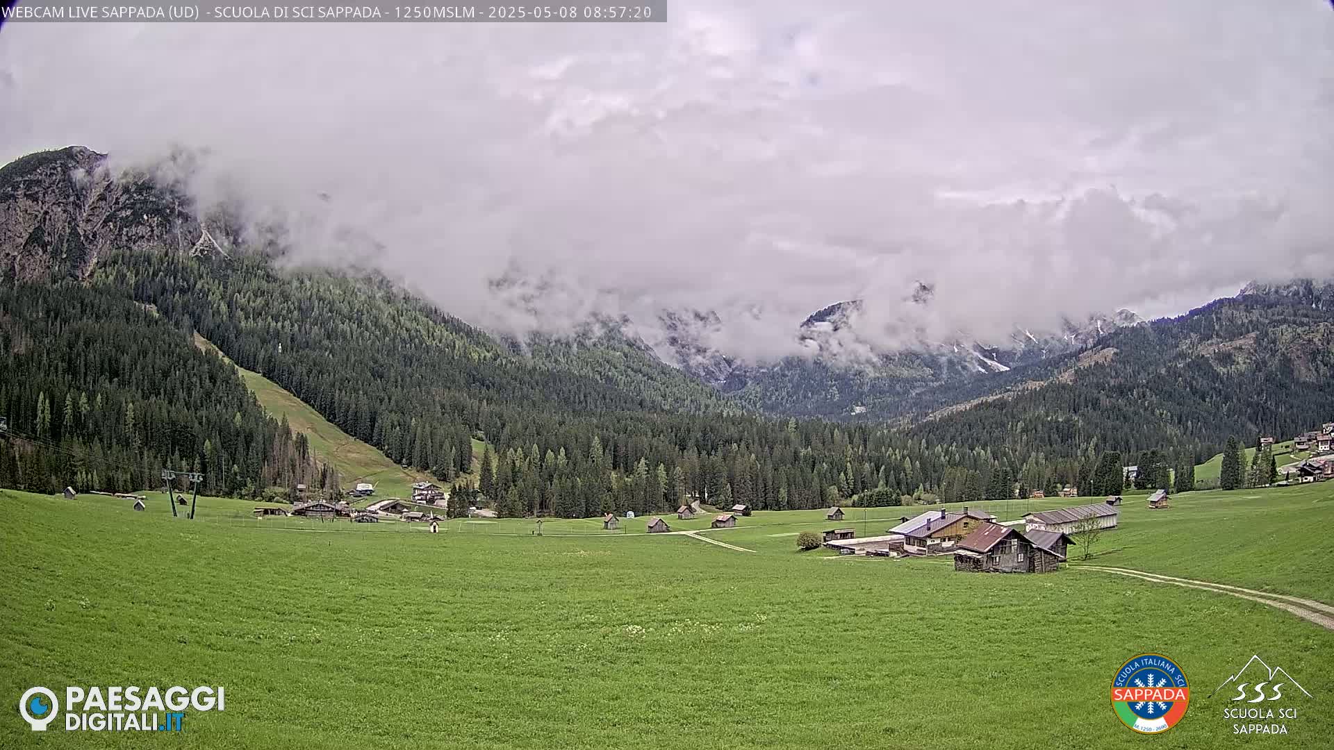 A valley of green fields and scattered buildings is nestled beneath cloud-covered mountains.