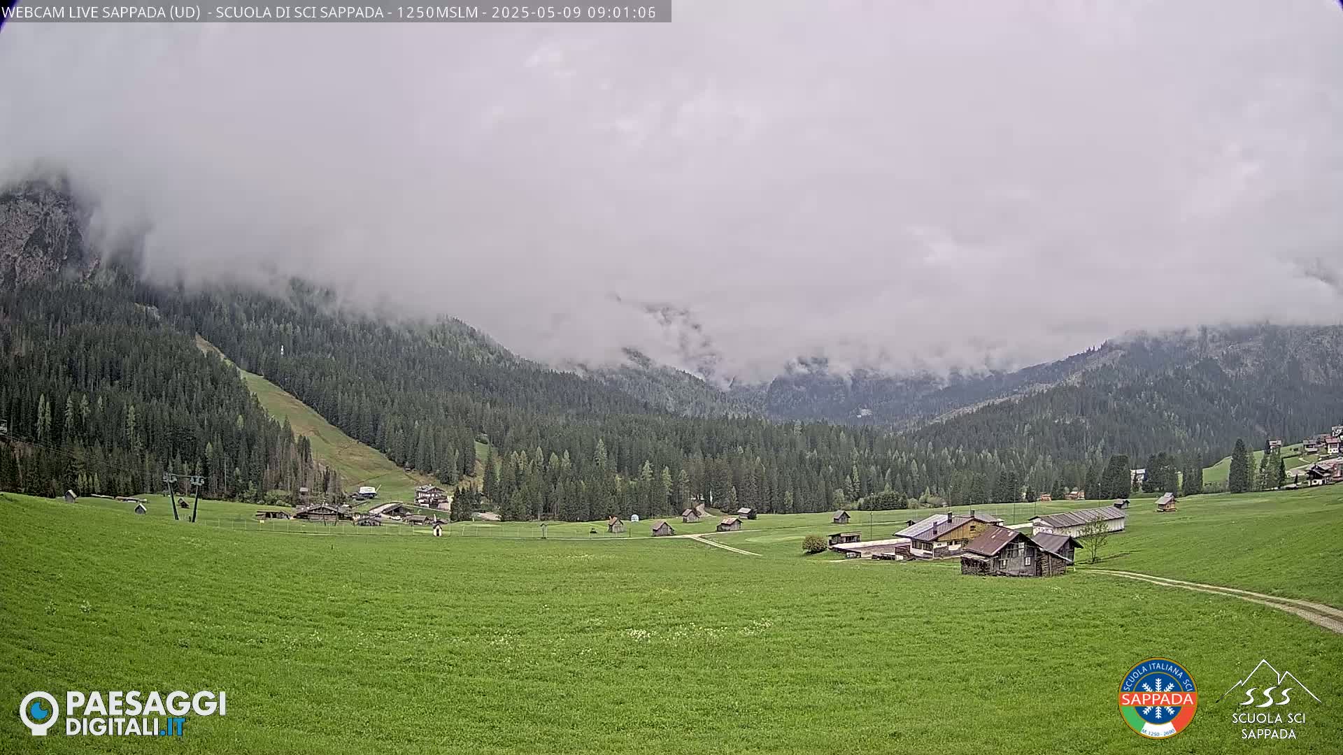 A valley of green fields and dark green coniferous forests is shrouded in a misty, overcast sky; a few small buildings are scattered throughout the valley.