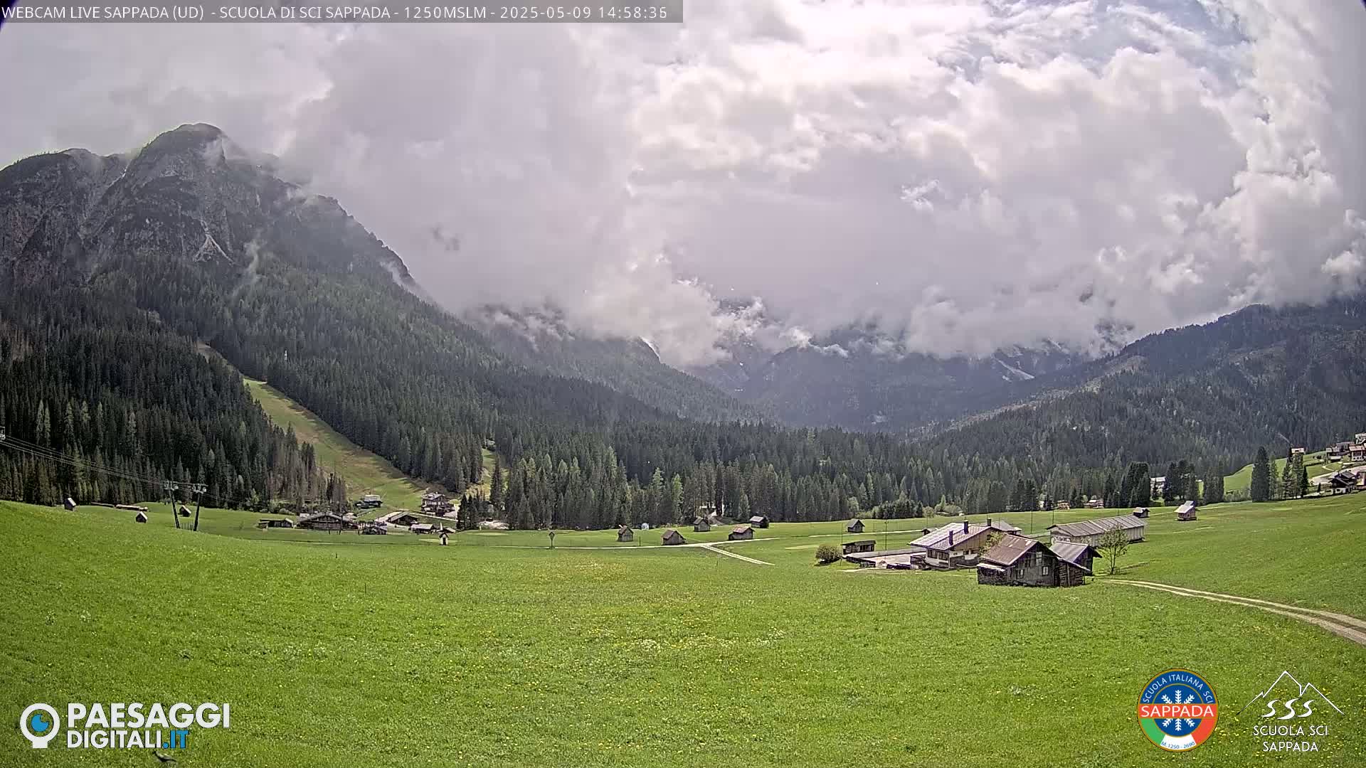 A partly cloudy day reveals a verdant valley dotted with small buildings, nestled amongst evergreen forests and mountains partially shrouded in mist.