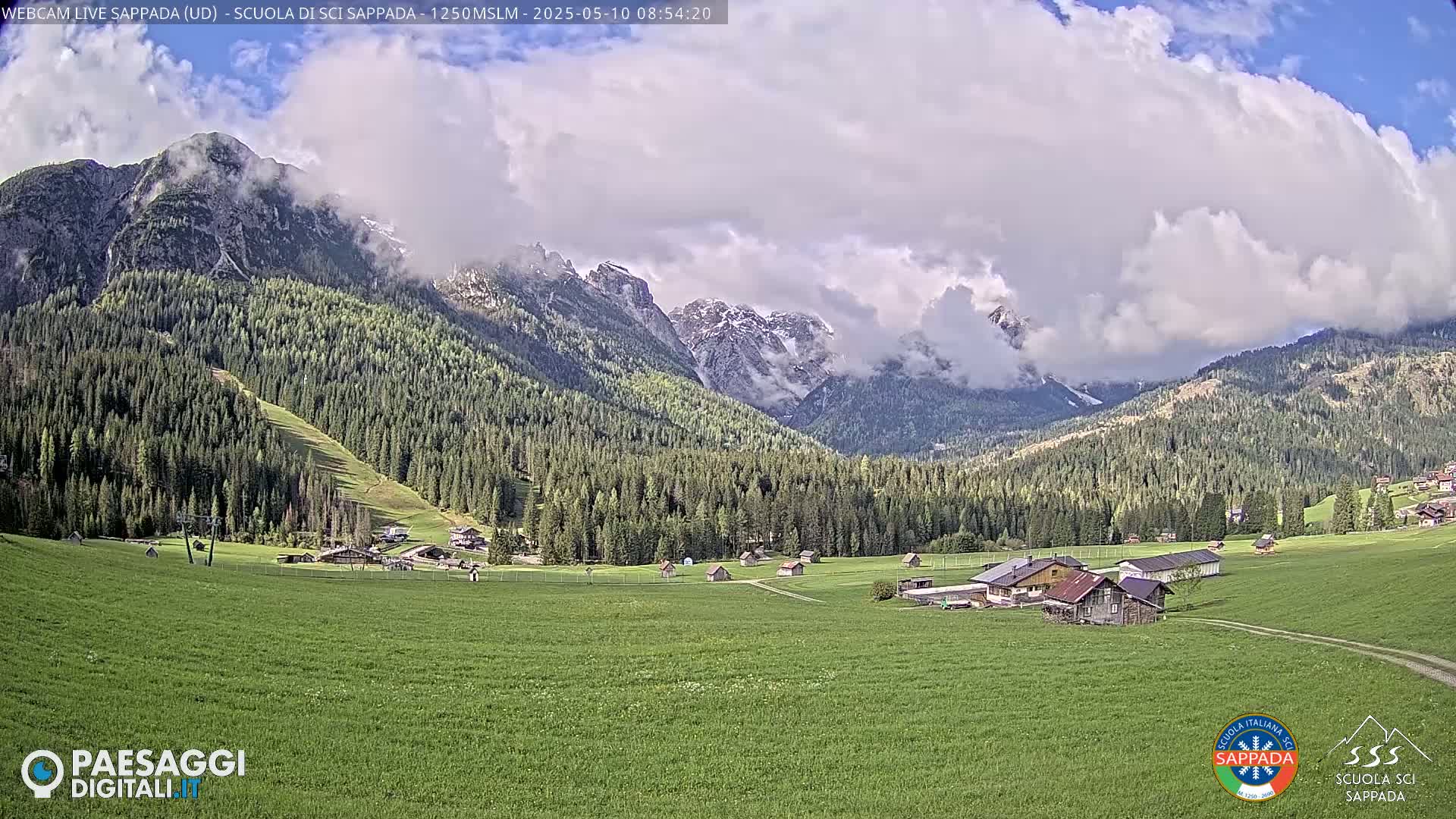 A partly cloudy day reveals a valley of green fields and small buildings nestled among a dark green forest, with snow-capped mountains in the background.