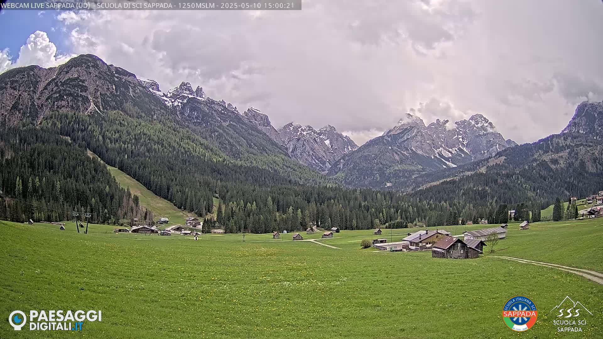 A verdant valley dotted with small buildings is nestled beneath snow-capped mountains under a cloudy sky.