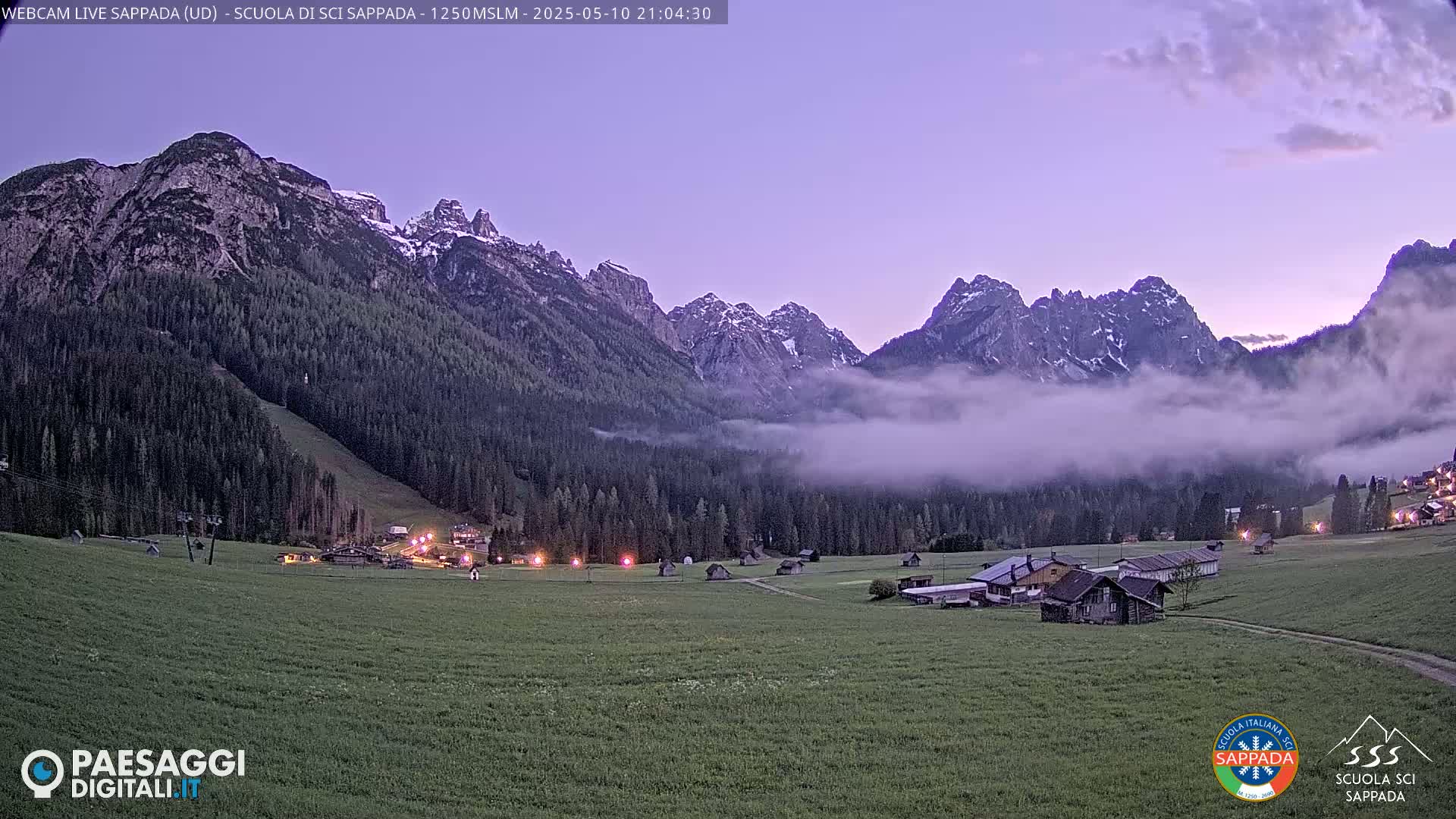 A valley at twilight, with low-lying fog partially obscuring snow-capped mountains in the background, is lit by lights from a small village in the foreground.