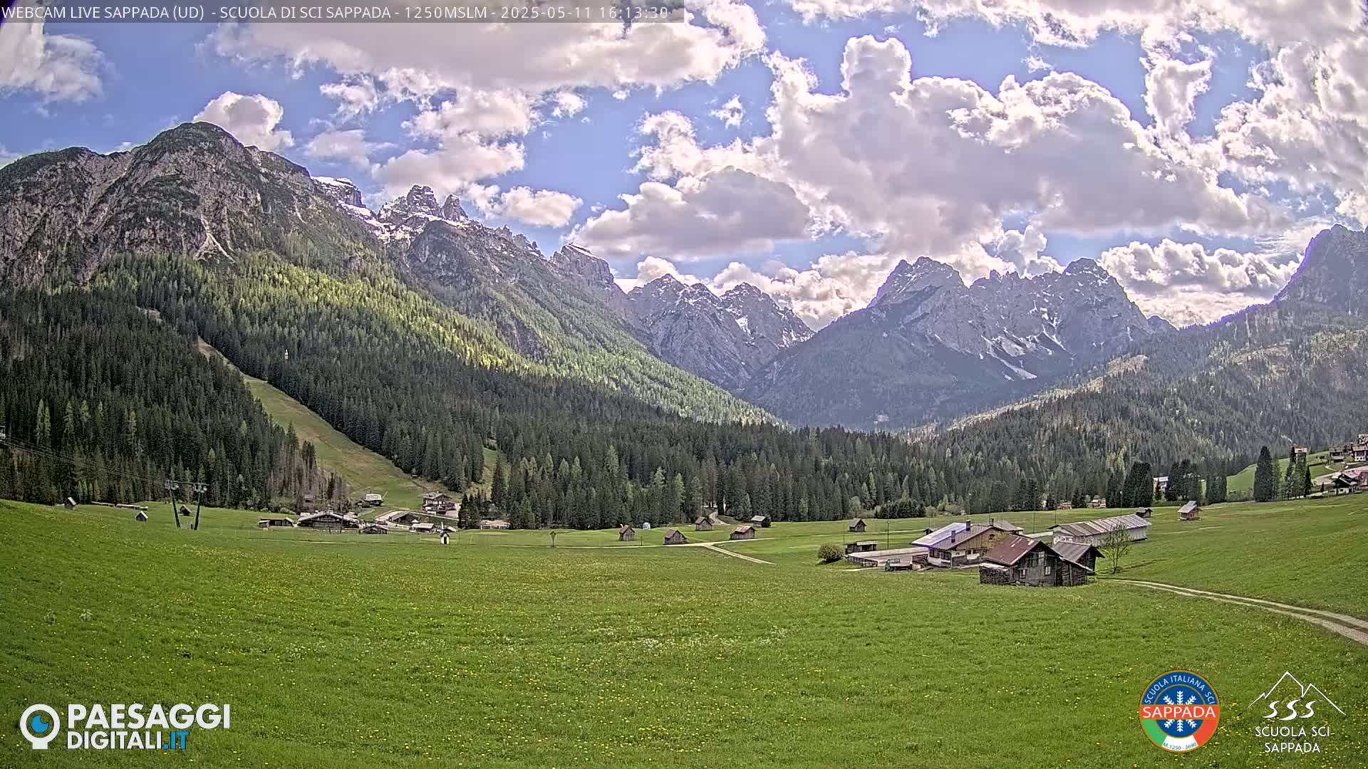 A partly cloudy day reveals a valley of green fields and small buildings nestled at the base of a mountain range partially covered in snow.