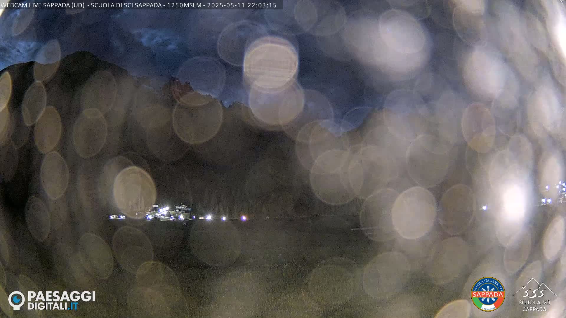 A nighttime view of a dark mountainside and a flat area with several lights in the distance, obscured by blurry, out-of-focus light spots.