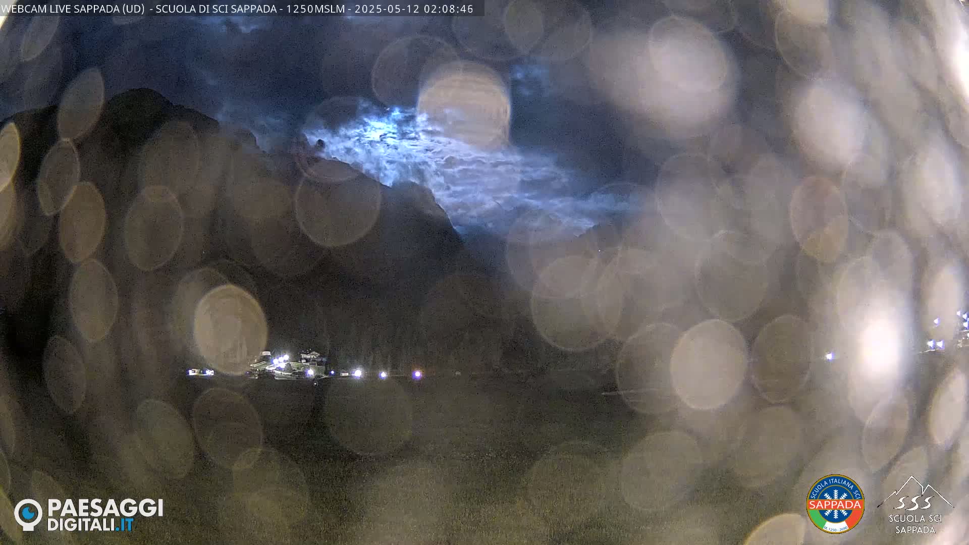 A nighttime view of a mountain range under a cloudy sky with a few lights visible in the foreground, obscured by raindrops or condensation on a window.