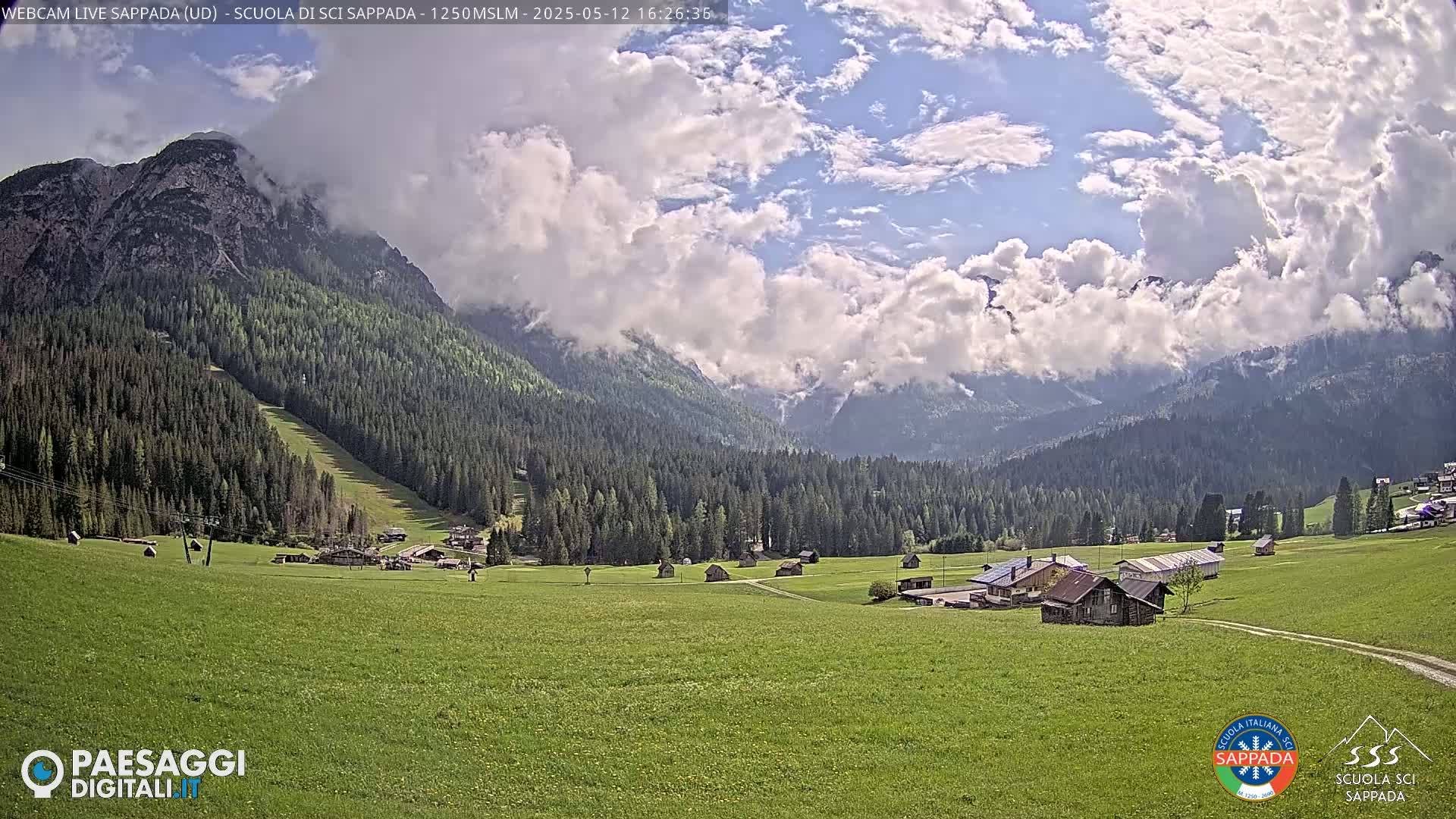 A partly cloudy sky overlooks a verdant valley with scattered buildings and a mountain in the background.