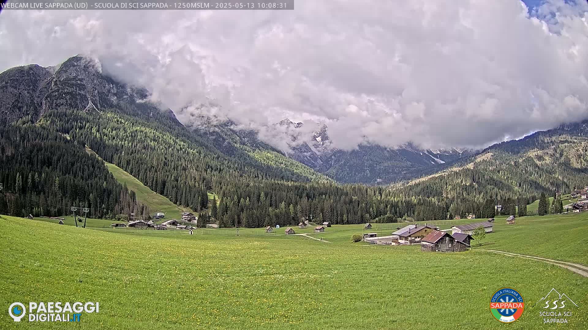 A partly cloudy mountain valley scene shows a village nestled amongst green fields and forests, with partially cloud-covered mountains in the background.