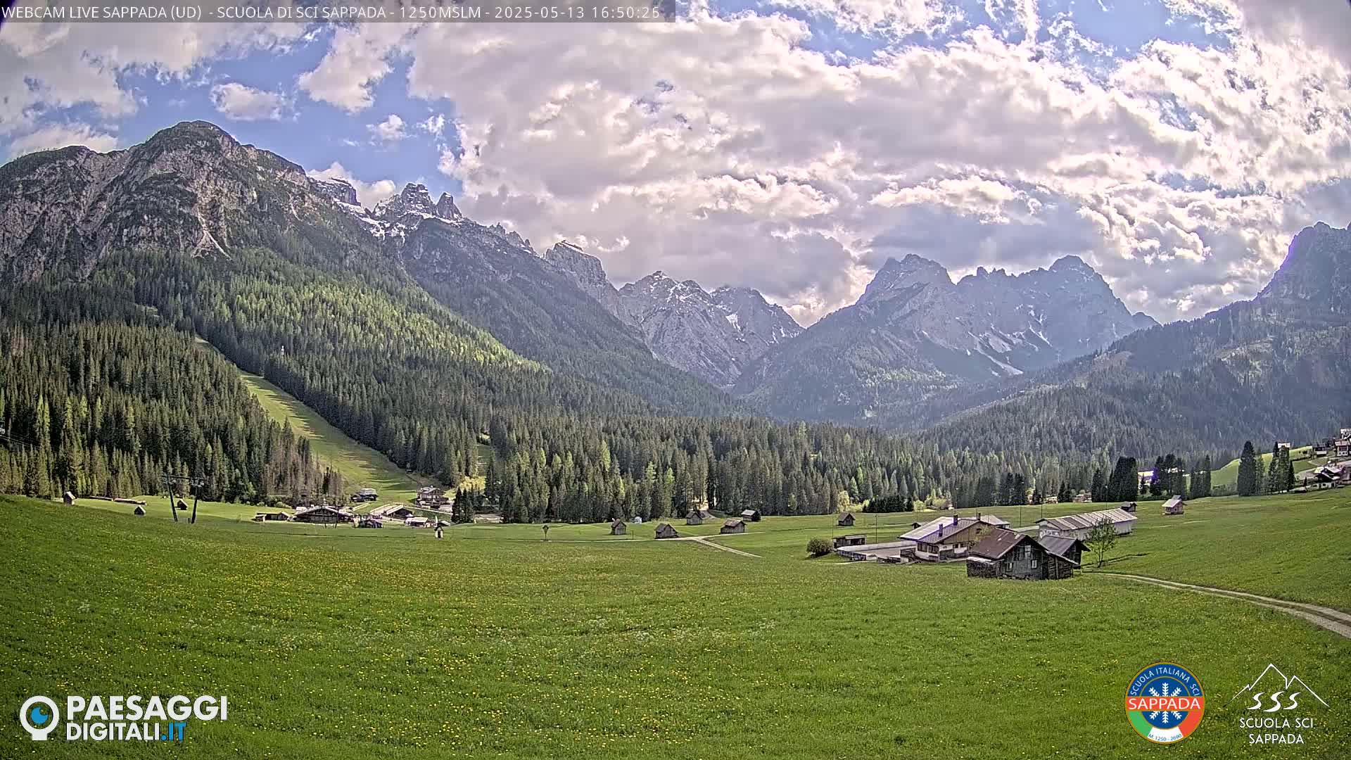 A partly cloudy day reveals a lush green valley dotted with small buildings, nestled between towering, snow-capped mountains covered in dark green forests.