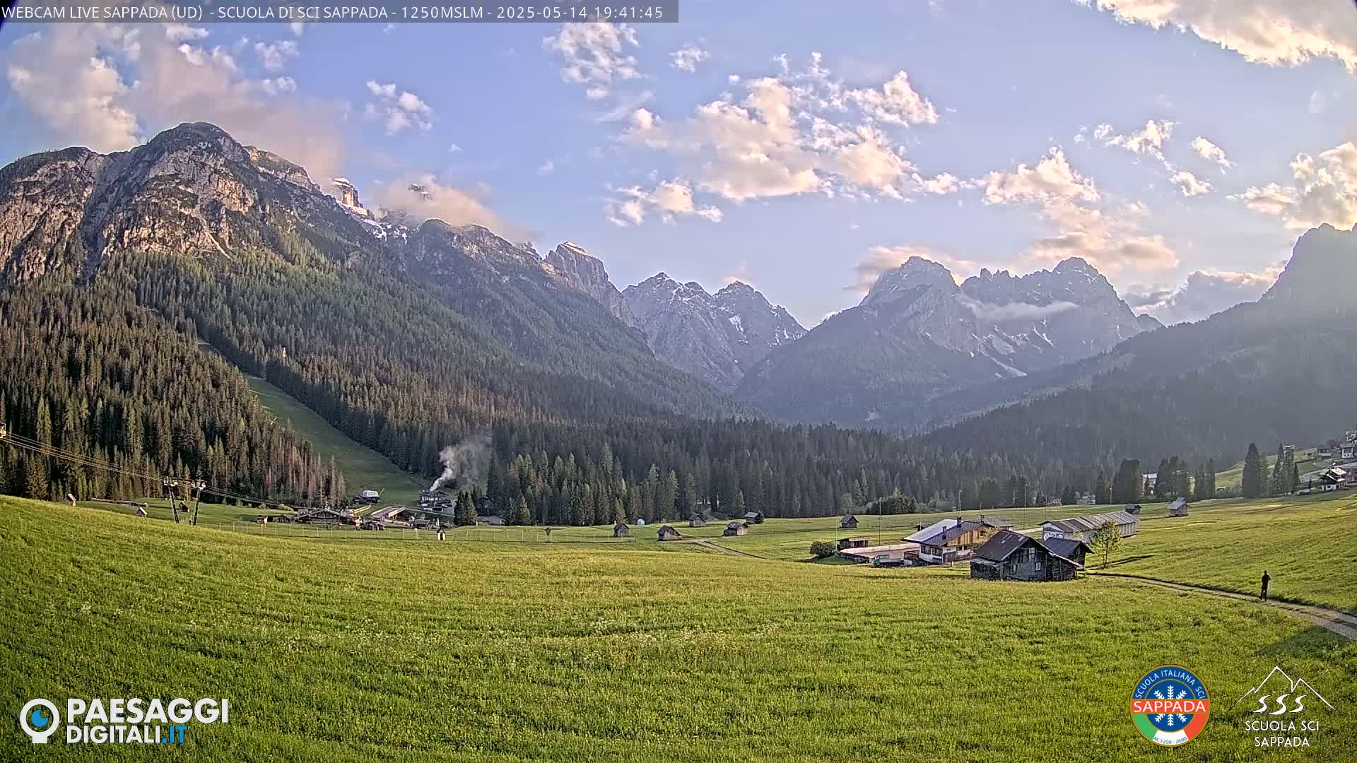 A partly cloudy sunset illuminates a valley of green fields and dark evergreen forests nestled between snow-capped mountains.