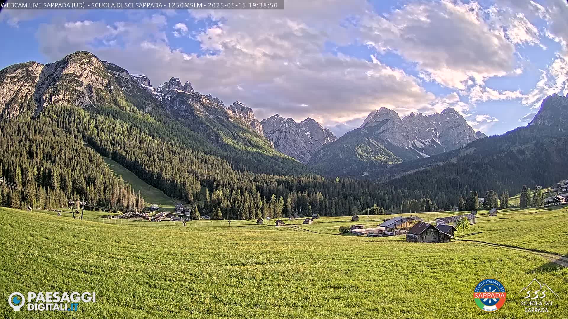 A valley of green fields and coniferous forests nestled beneath a mountain range with partially cloudy skies.